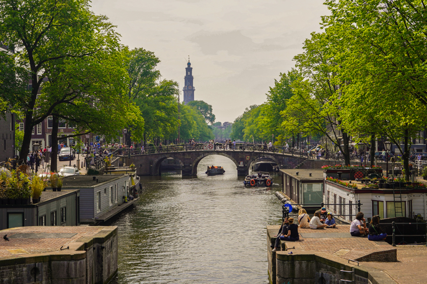 A classic Amsterdam canal view with the Westerkerk in the distance, lively bridges, houseboats and people enjoying the waterfront, shot by Louise Kluit