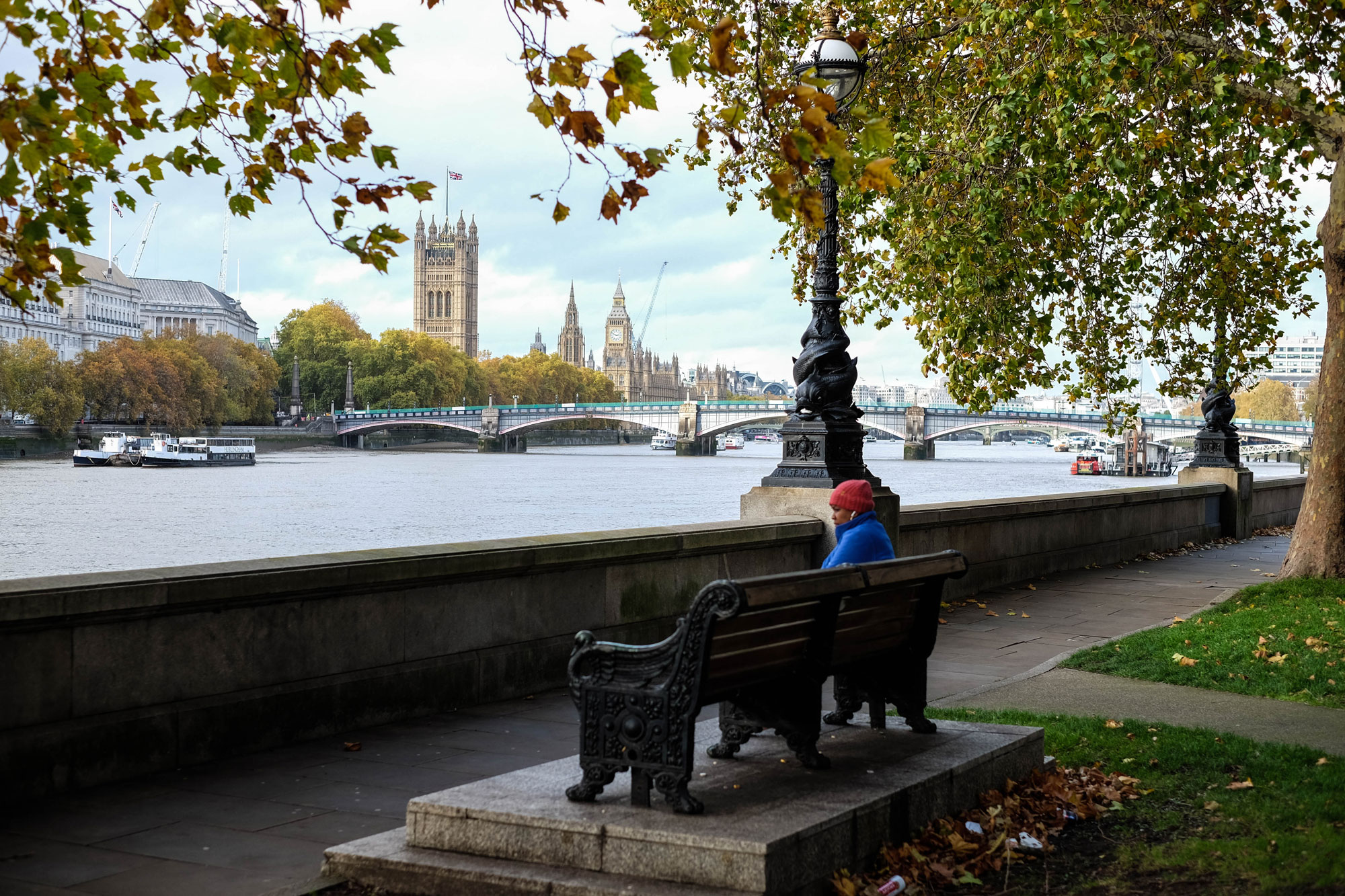 A person sits on a bench on the bank of the River Thames in London, England. The Houses of Parliament are visible in the background, with a Union Jack flag flying above. The person is wearing a blue coat and a red hat, matching the colours of the flag.