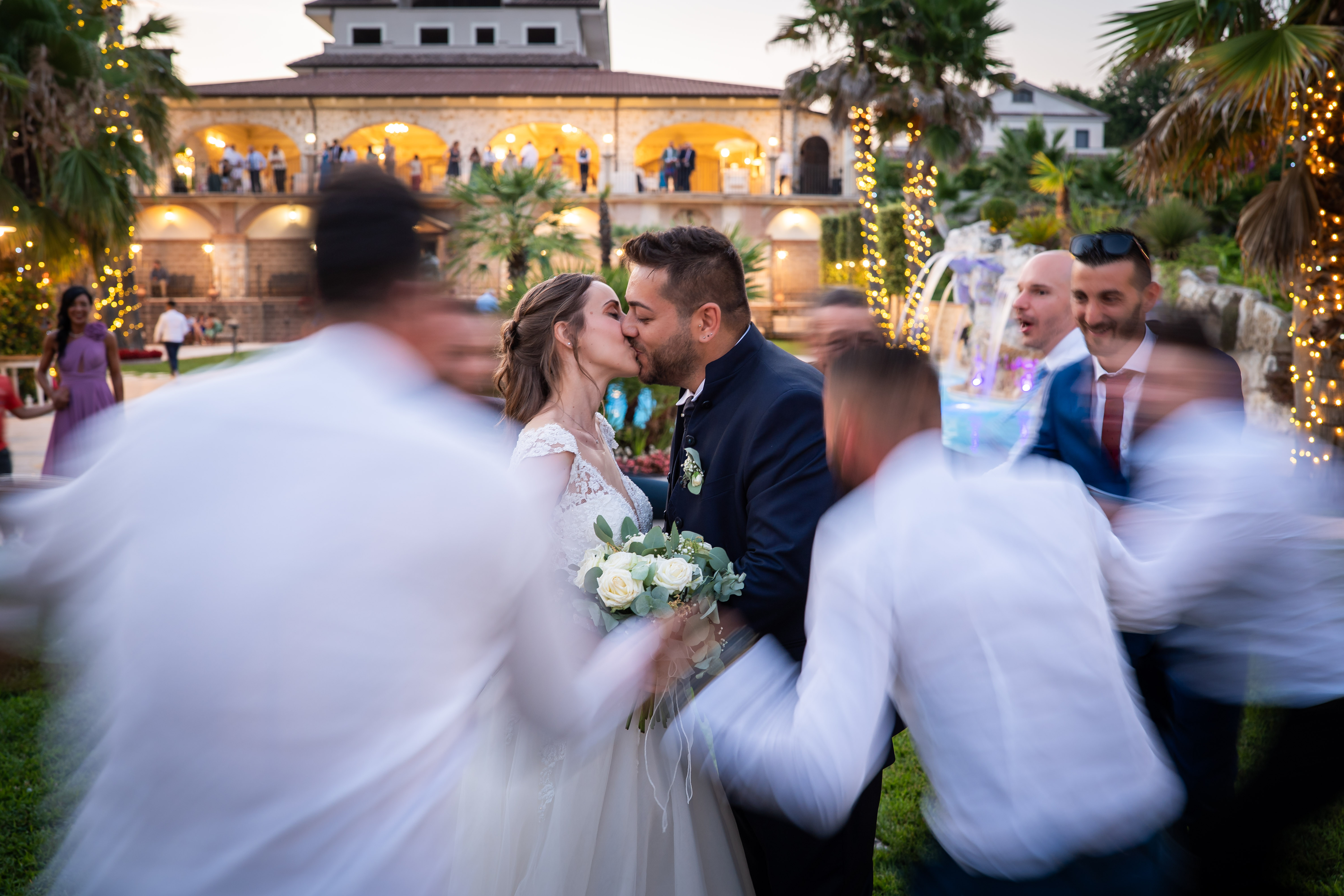 Sposi che si baciano nitidi al centro, mentre gli invitati intorno appaiono sfocati dal movimento, in un giardino illuminato.