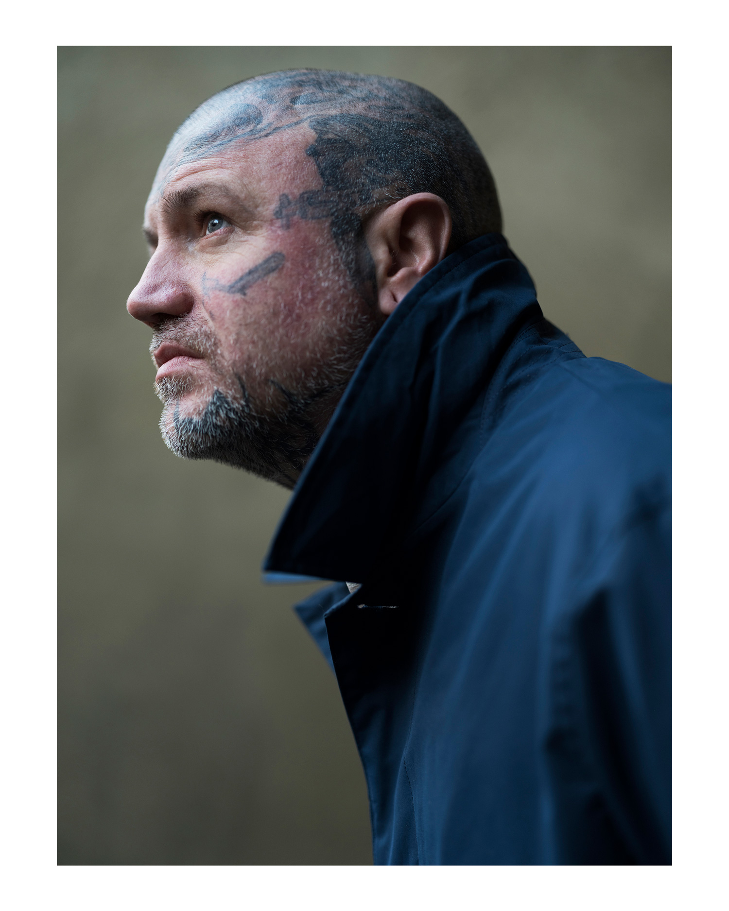 Nahaufnahme eines tätowierten Mannes in einem blauen Mantel, der vor einer Wand steht, fotografiert von Simon Murphy mit einer Fujifilm GFX 50S.