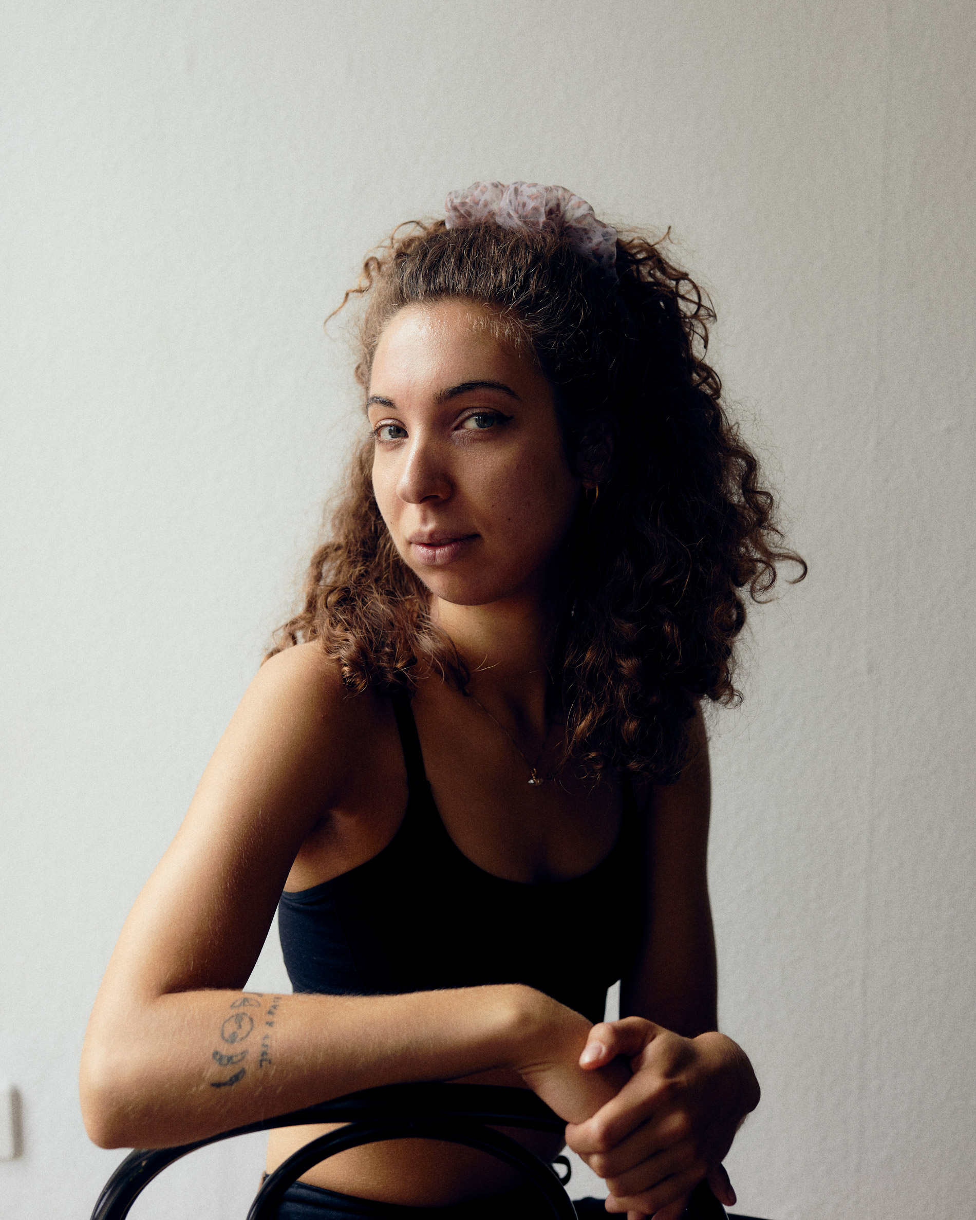 Porträt einer Person, die auf einem Stuhl sitzt mit Händen auf der Lehne und in die Kamera blickend

