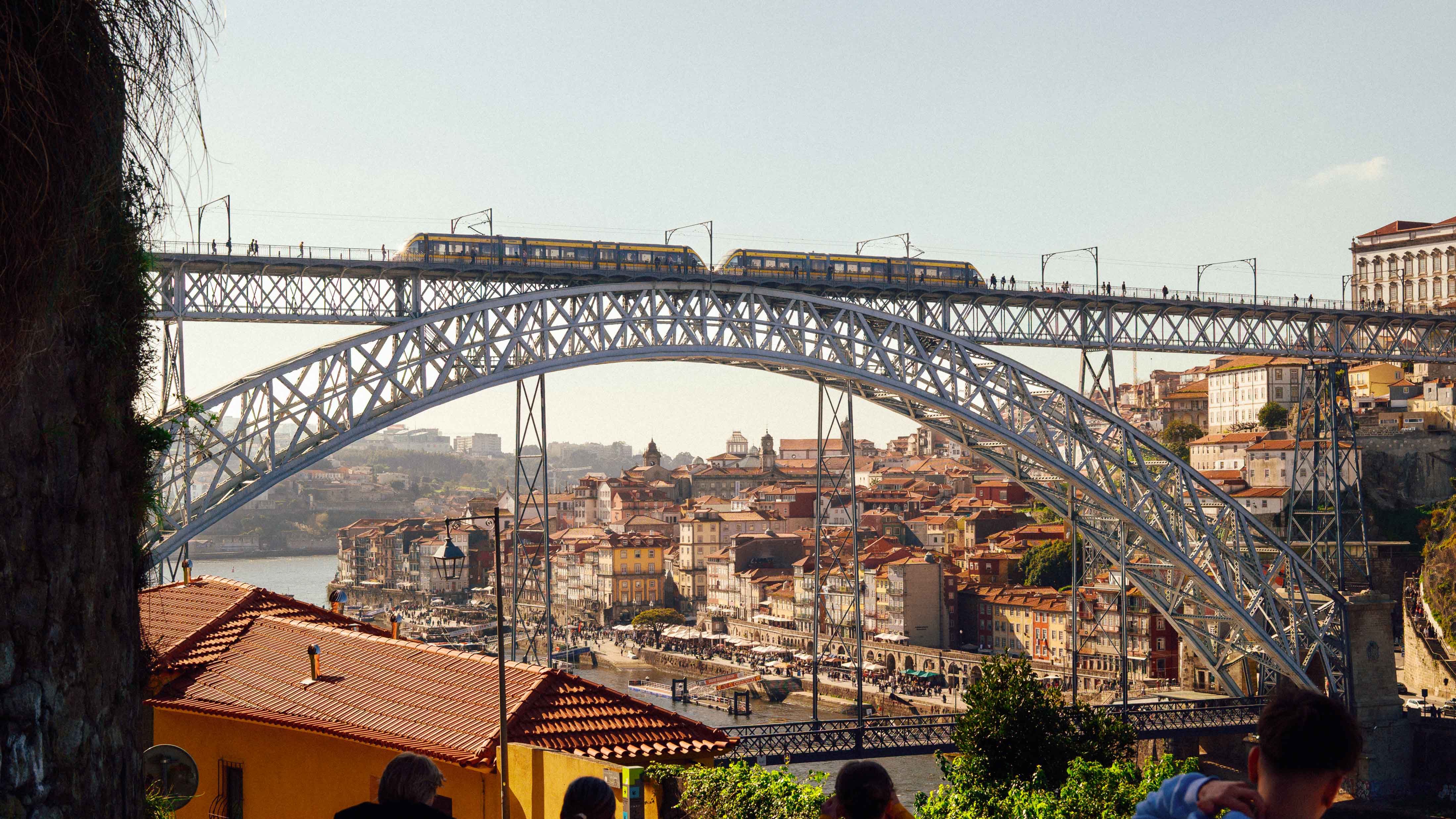 Dom Luís I Bridge spanning the Douro River with a metro train crossing the upper deck and Porto’s Ribeira district rising in layered rooftops beneath the arch.
