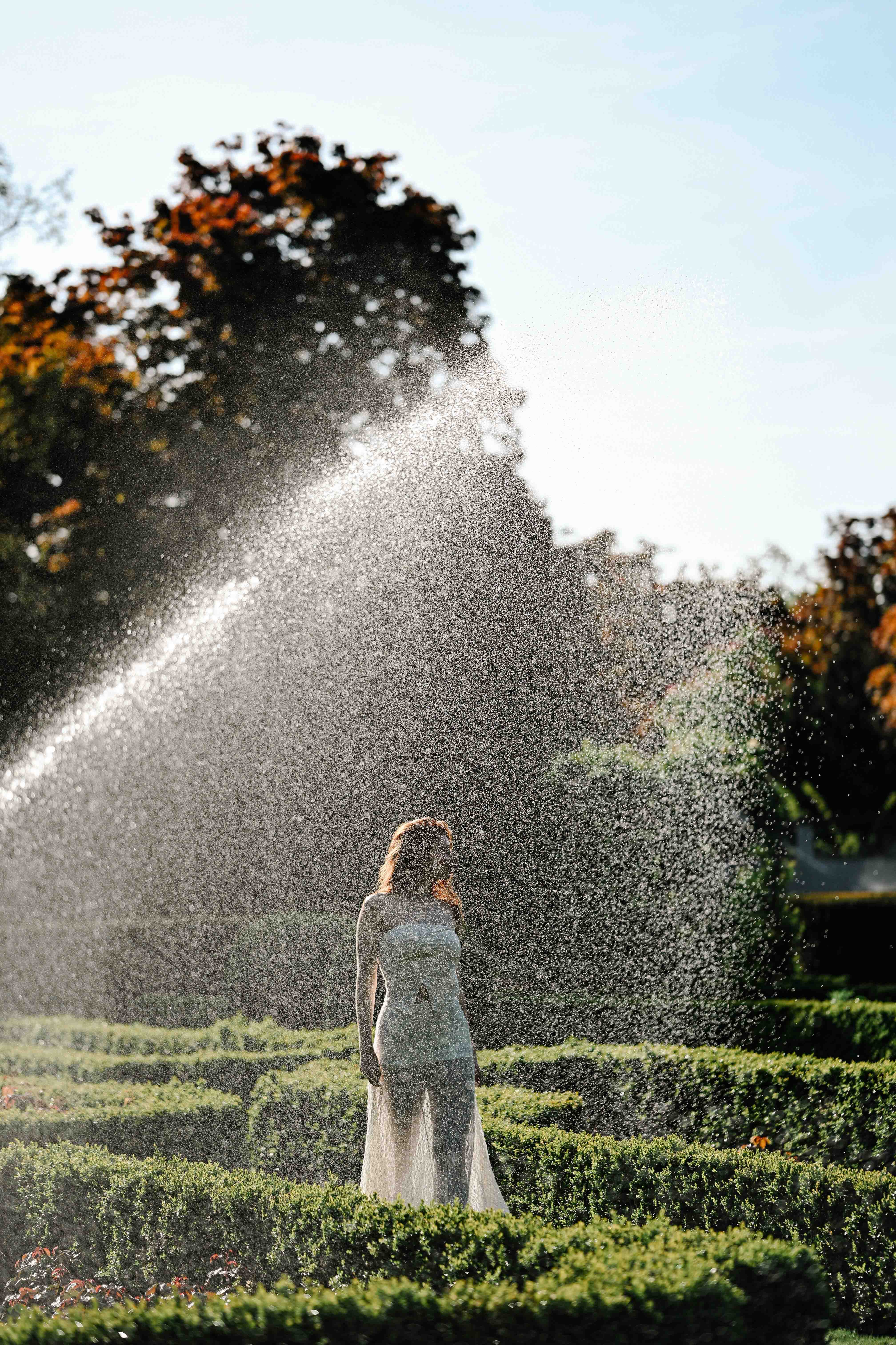 A model standing under a garden sprinkler, wearing a white dress on a sunny morning at Berlin’s Humboldthain park. Shot by Alina Rudya on a Nikon Z6 III.