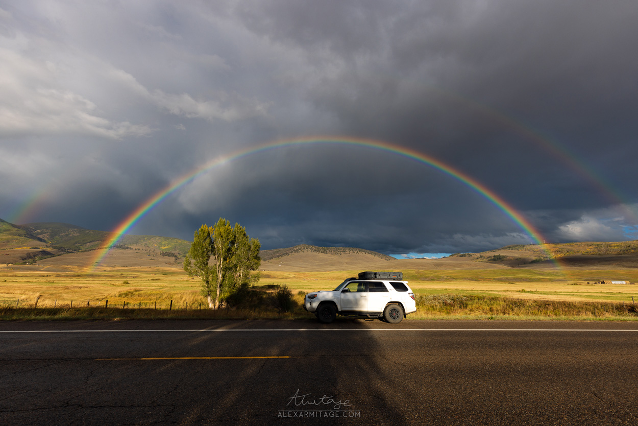 A vehicle sits parked by the road with a rainbow in the background.
