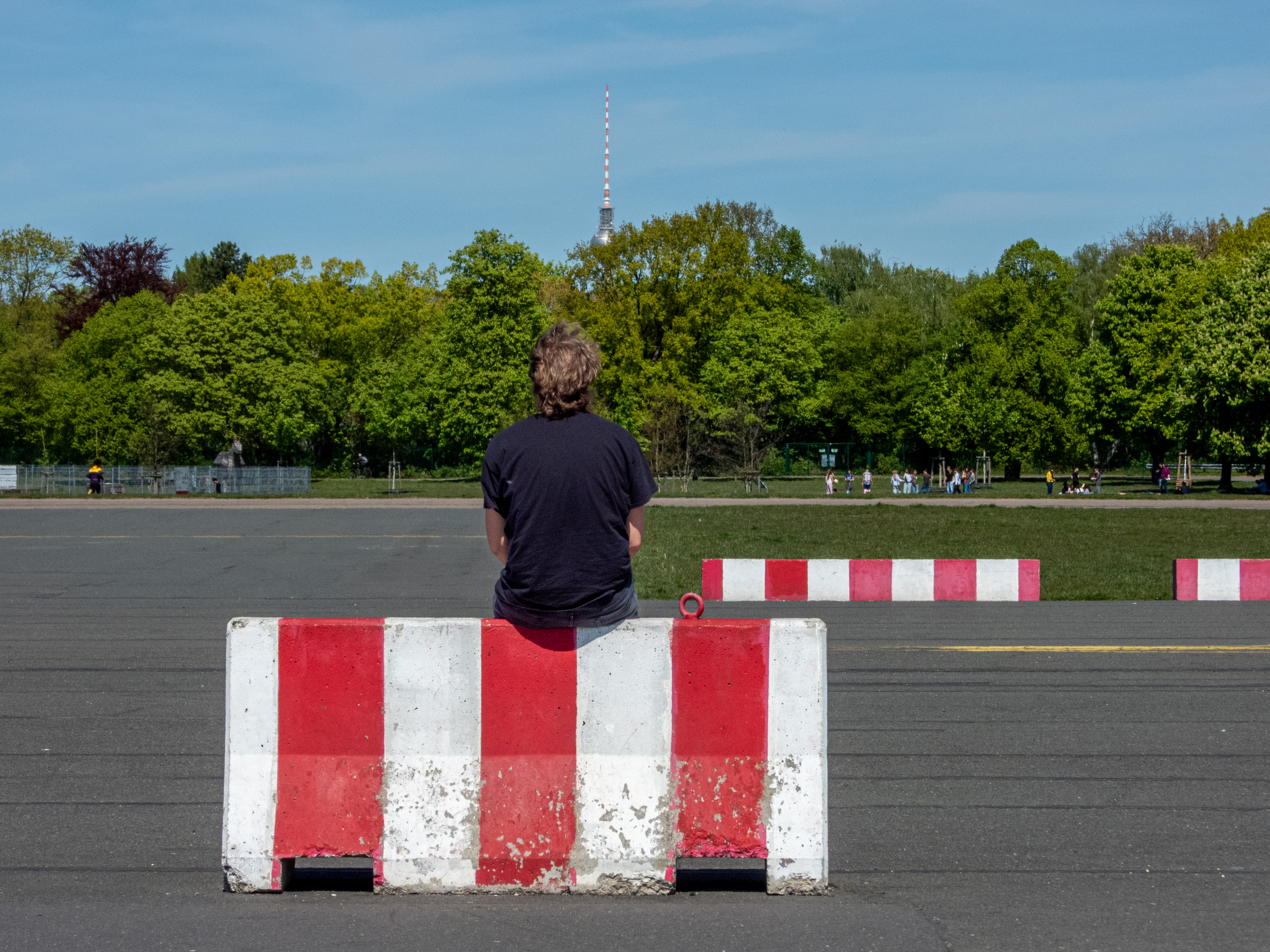 Una persona sentada en una barrera en el Parque Tempelhof de Berlín, con la torre de televisión de fondo.