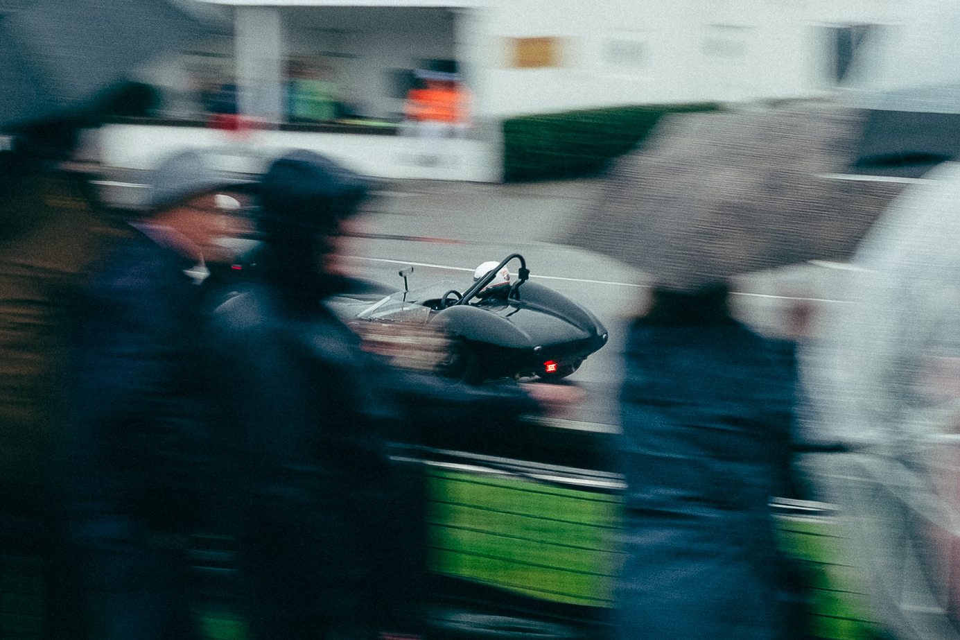 A dark racing green vintage car going fast on a racing track with spectators blurred out in the foreground. Taken by Connor Redmond on the Fujifilm X100F.