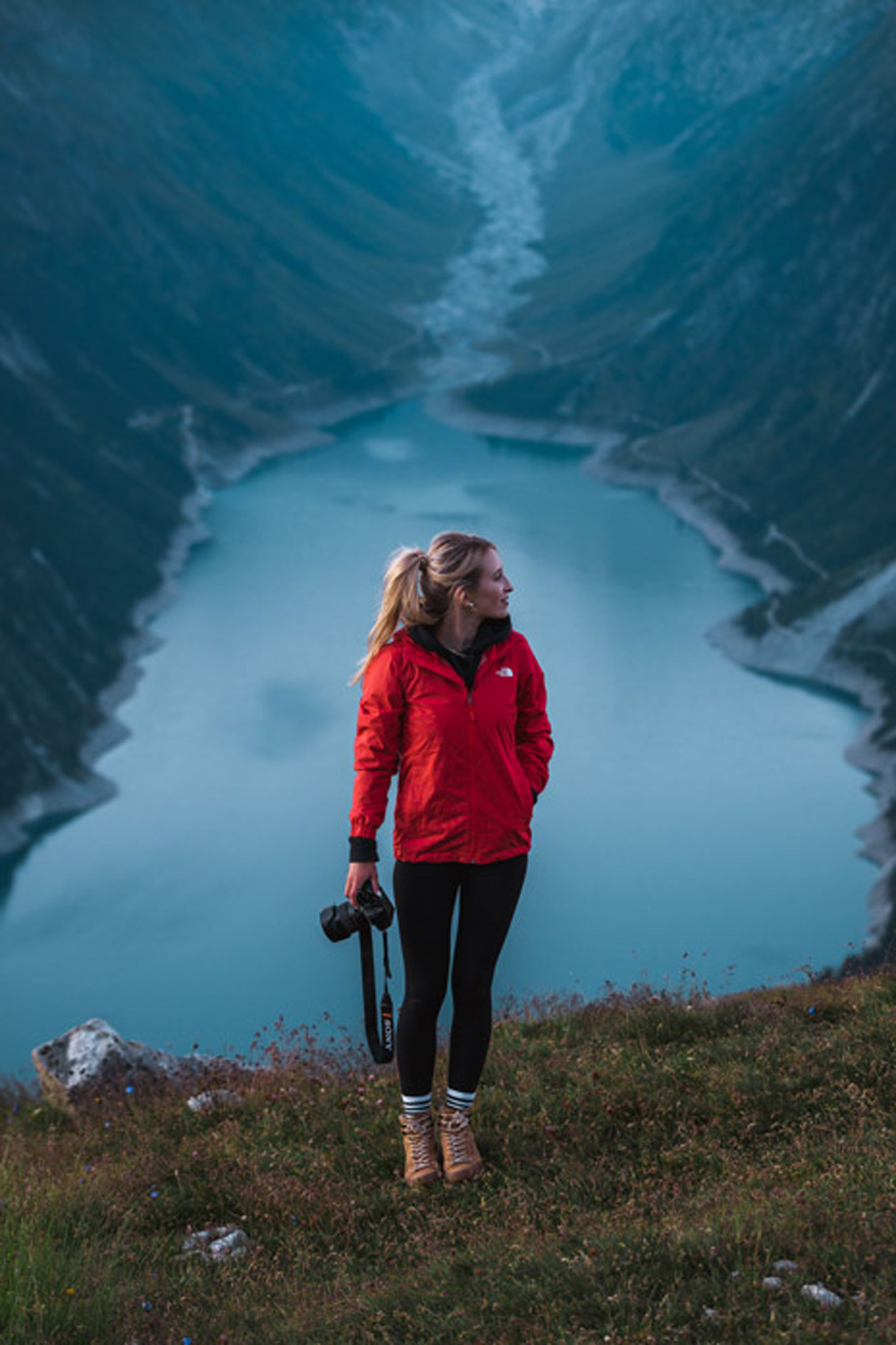 Bild von einer Frau mit Kamera in der Hand, die nach links blickt und vor einem Bergsee steht.
