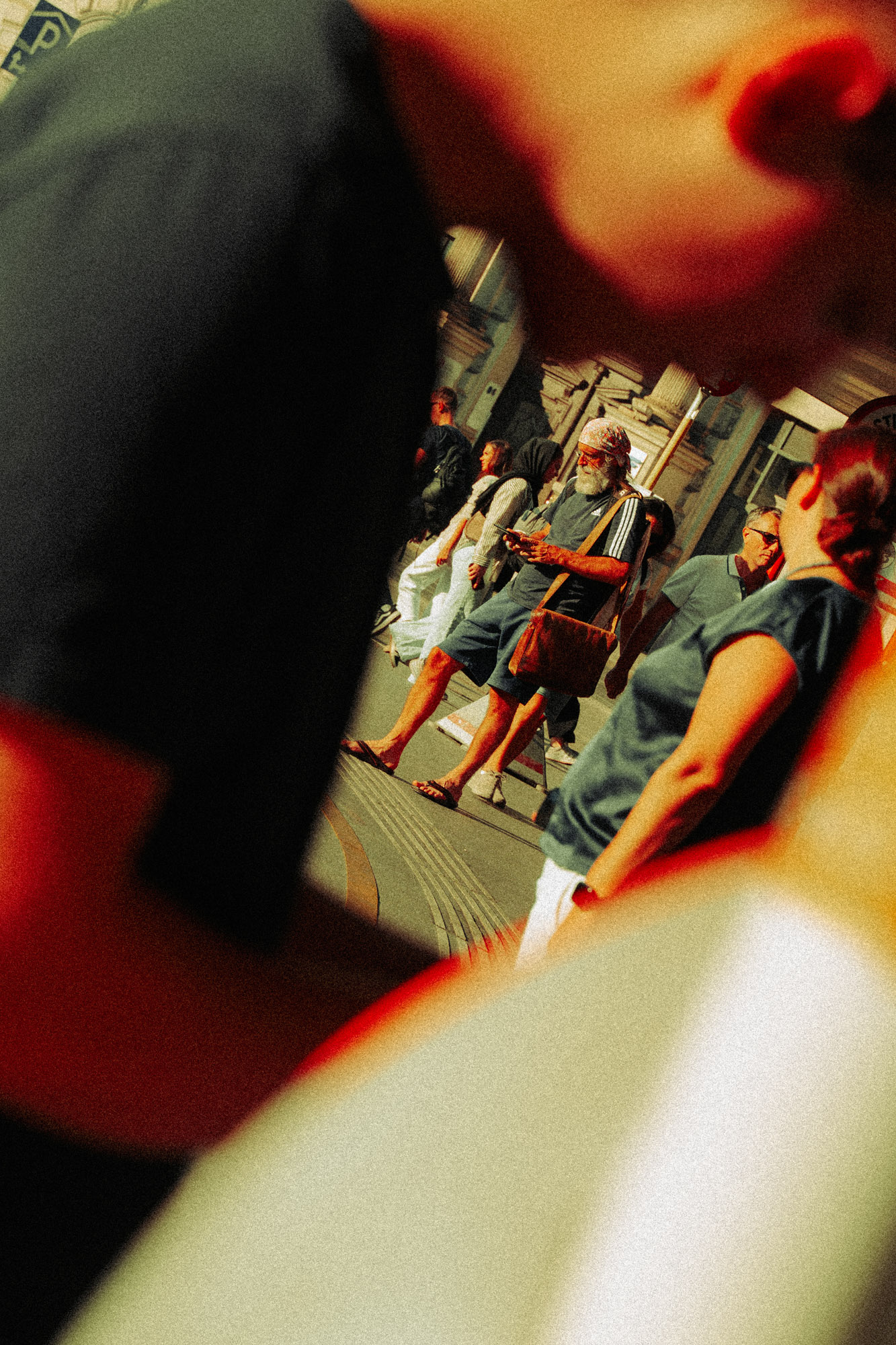 Streetfoto einer schräg komponierten Szene in Wien, in der mehrere Passant:innen und Passanten im Hintergrund zwischen unscharfen Elementen im Vordergrund zu sehen sind, was dem Bild eine dichte, beobachtende Wirkung verleiht. Aufgenommen von Dhan Limwattana mit einer Nikon Z5 II.