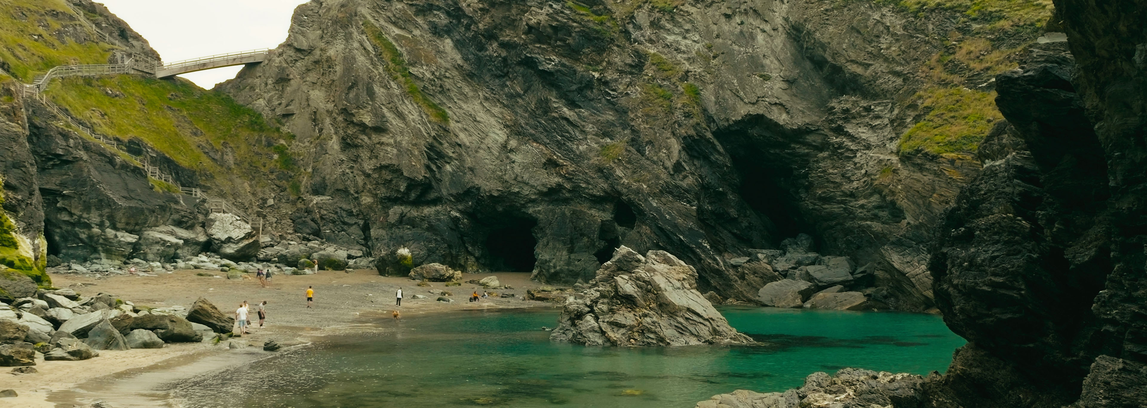 Coastal cove with rocky cliffs, sea caves and shallow turquoise water, with people exploring the beach below.