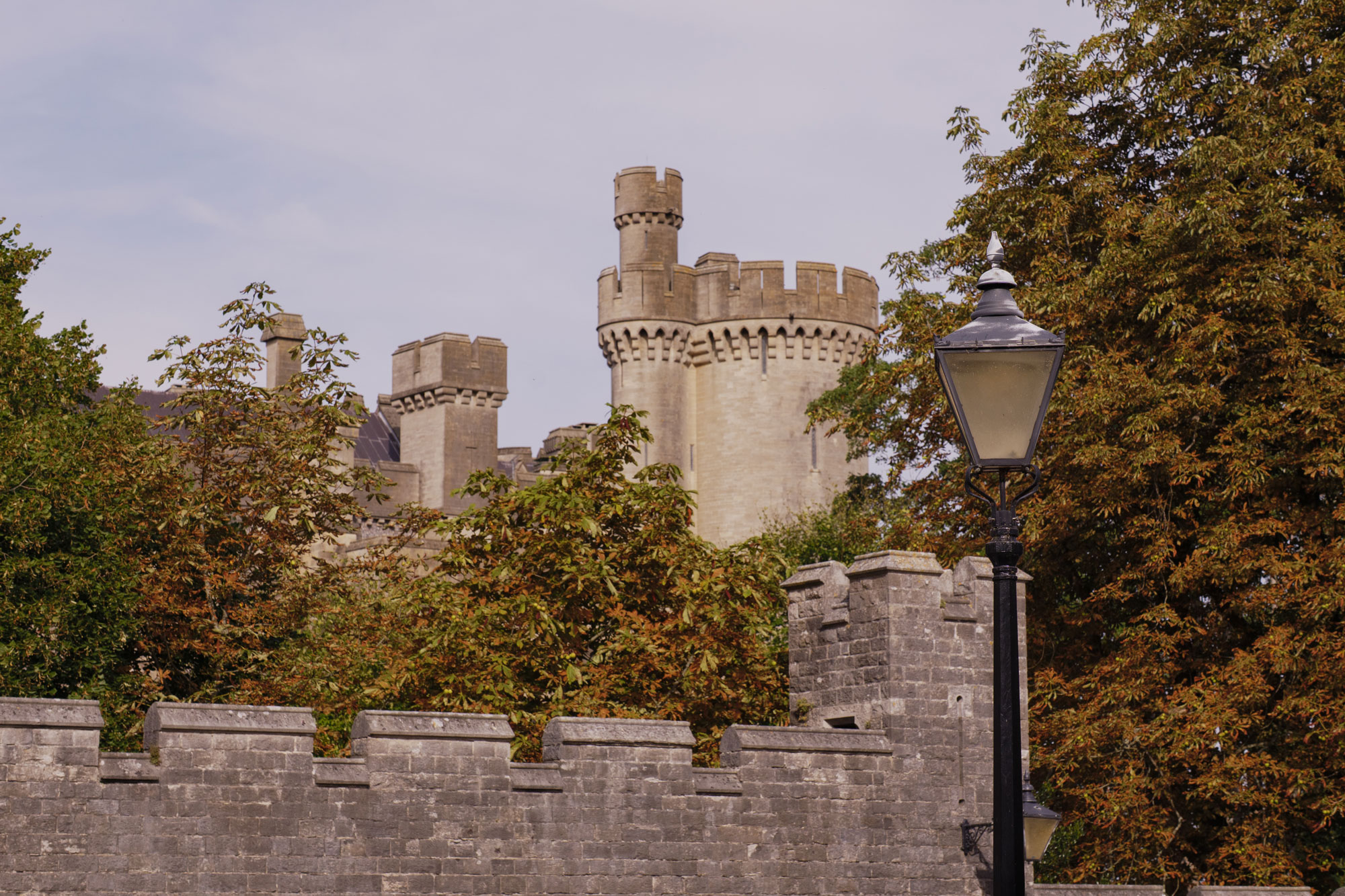 A castle wall, with its turrets in the background on a cloudy day. An old, ornate black street lamp, surrounded by trees, is in the foreground.