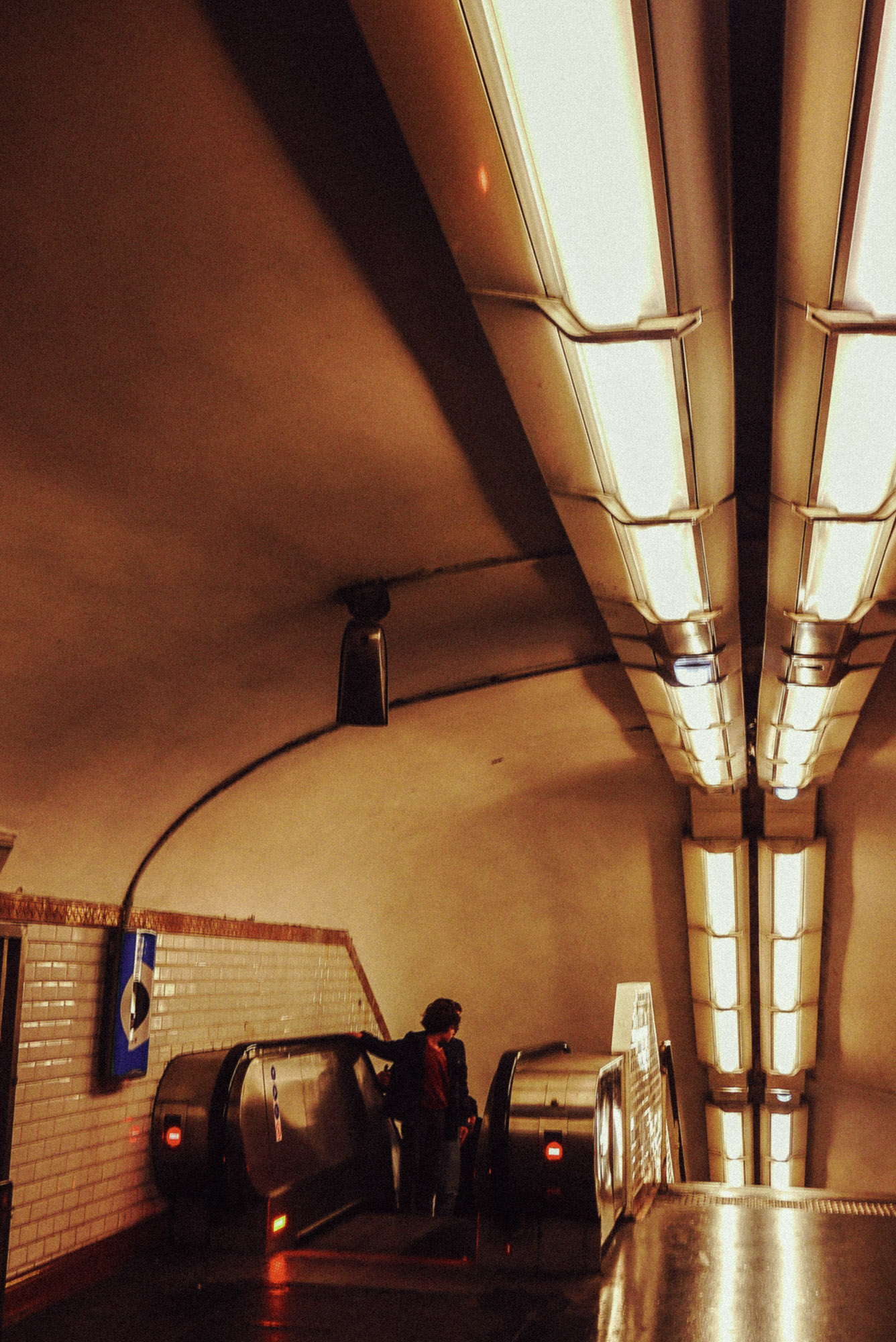 Streetaufnahme mit einer Person, die eine Rolltreppe in einer Pariser Métro-Station hinunterfährt. Links sind geflieste Wände und darüber lange, fluoreszierende Deckenleuchten zu sehen, die im Untergrund für Tiefe und Symmetrie sorgen. Aufgenommen von Wesley Verhoeve mit einer Panasonic Lumix GF1.