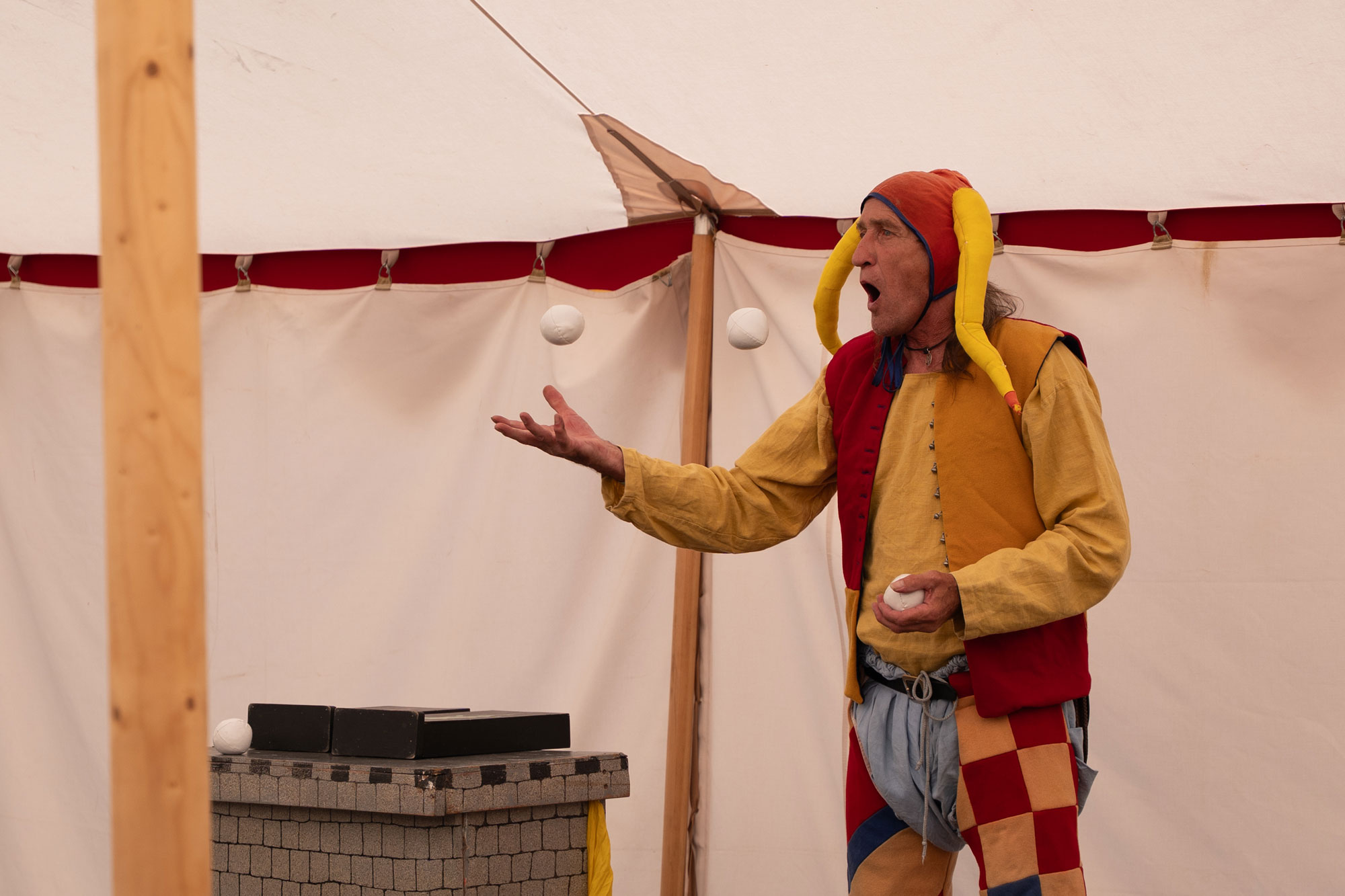 A person under a bell tent in a red and yellow jester costume. Their mouth is open and they are juggling two white balls.