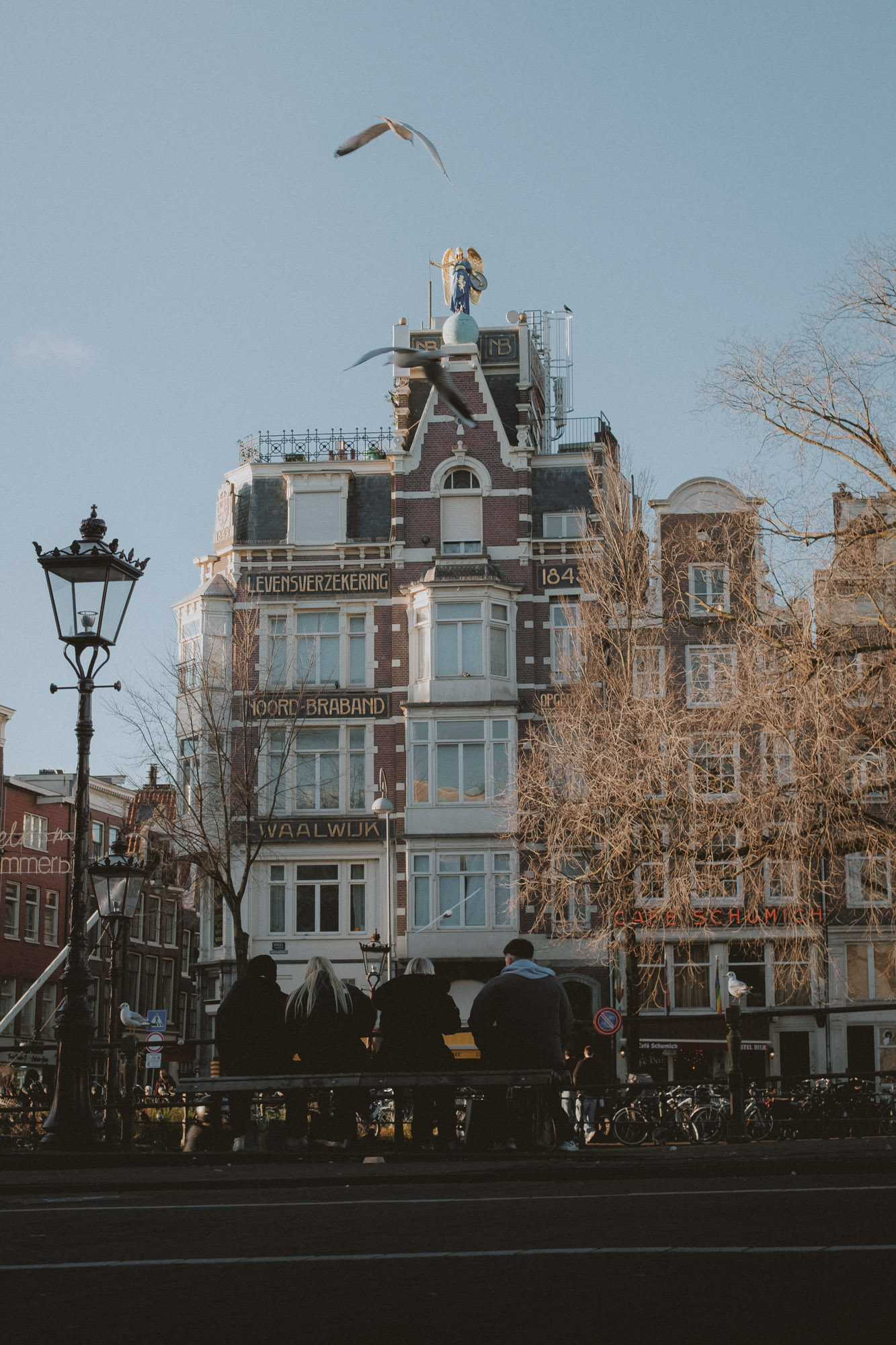 Bâtiment historique d’Amsterdam avec façade ornée et statue sur le toit, mouettes en vol et passants longeant le canal dans une lumière chaude de fin de journée
