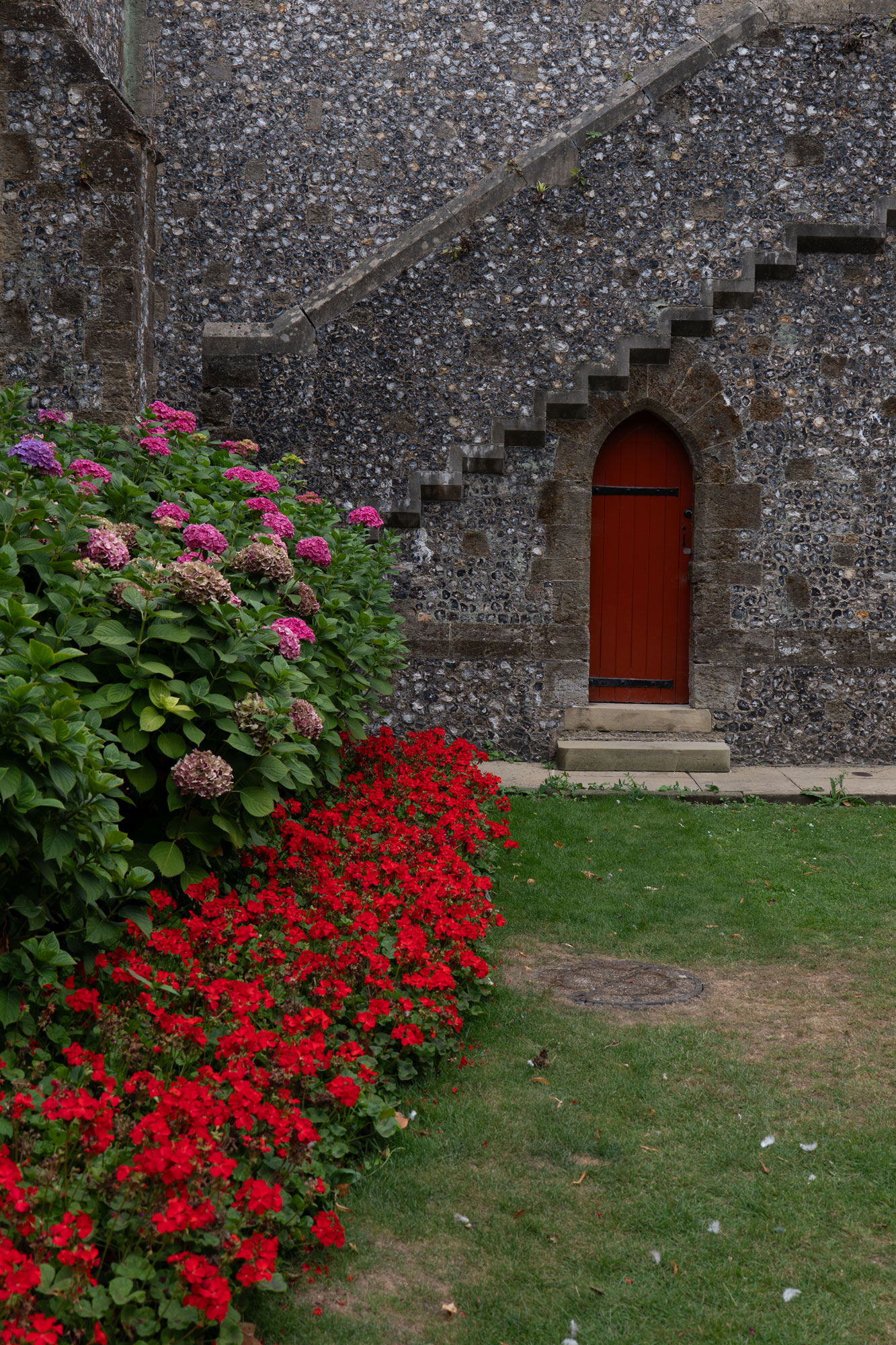 A wooden red door on the side of an old stone building, with an ornate staircase above. In the foreground, a grassy lawn is lined with a bed of red flowers. Above that, there are some Hydrangea bushes with pink flowers.