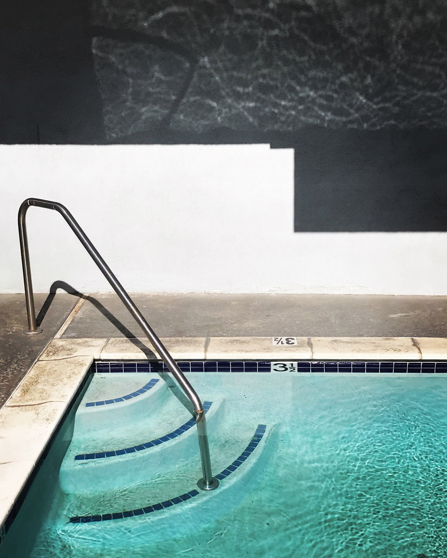 A staircase leads down into a swimming pool. There are three white loungers in the background, and a stick floating in the water. Shot by Kim Fuller.