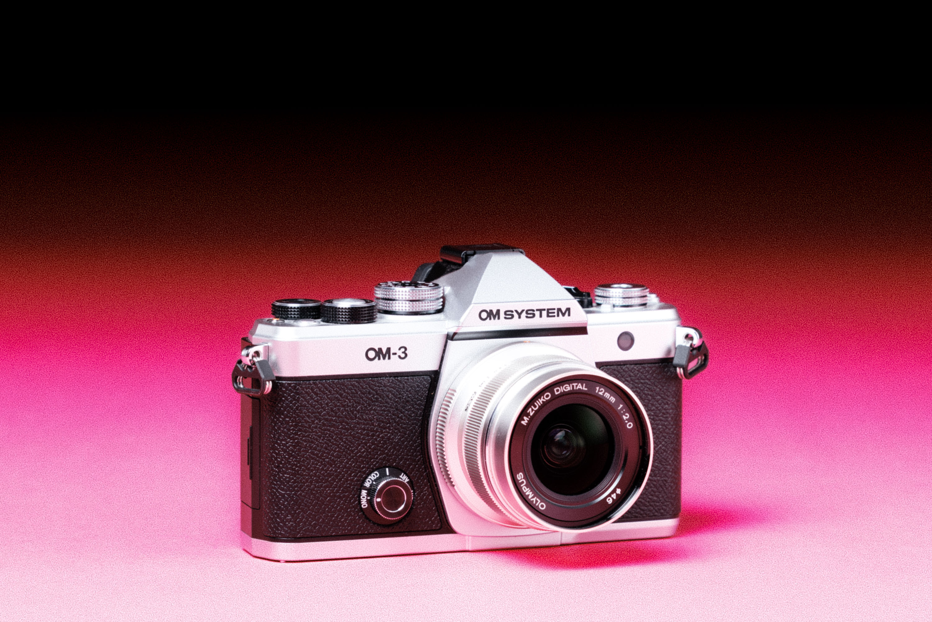 A used OM System OM-3 with an Olympus M.Zuiko Digital ED 12mm f/2.0 lens, with an added film-like grain effect on a graduated pink background.