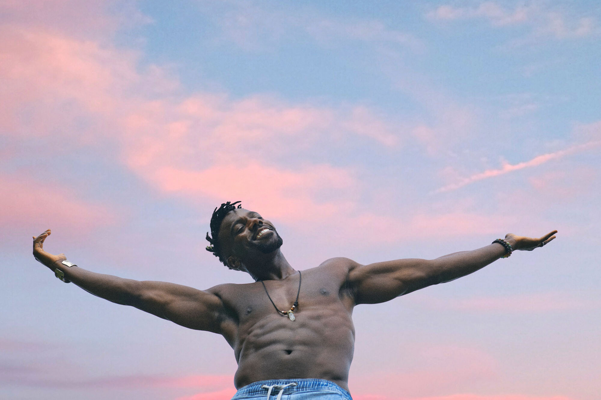 A low-angle portrait of a model wearing a pendant and blue shorts smiling and raising their arms outstretched with a blue-pink sky overhead, looking happy and content