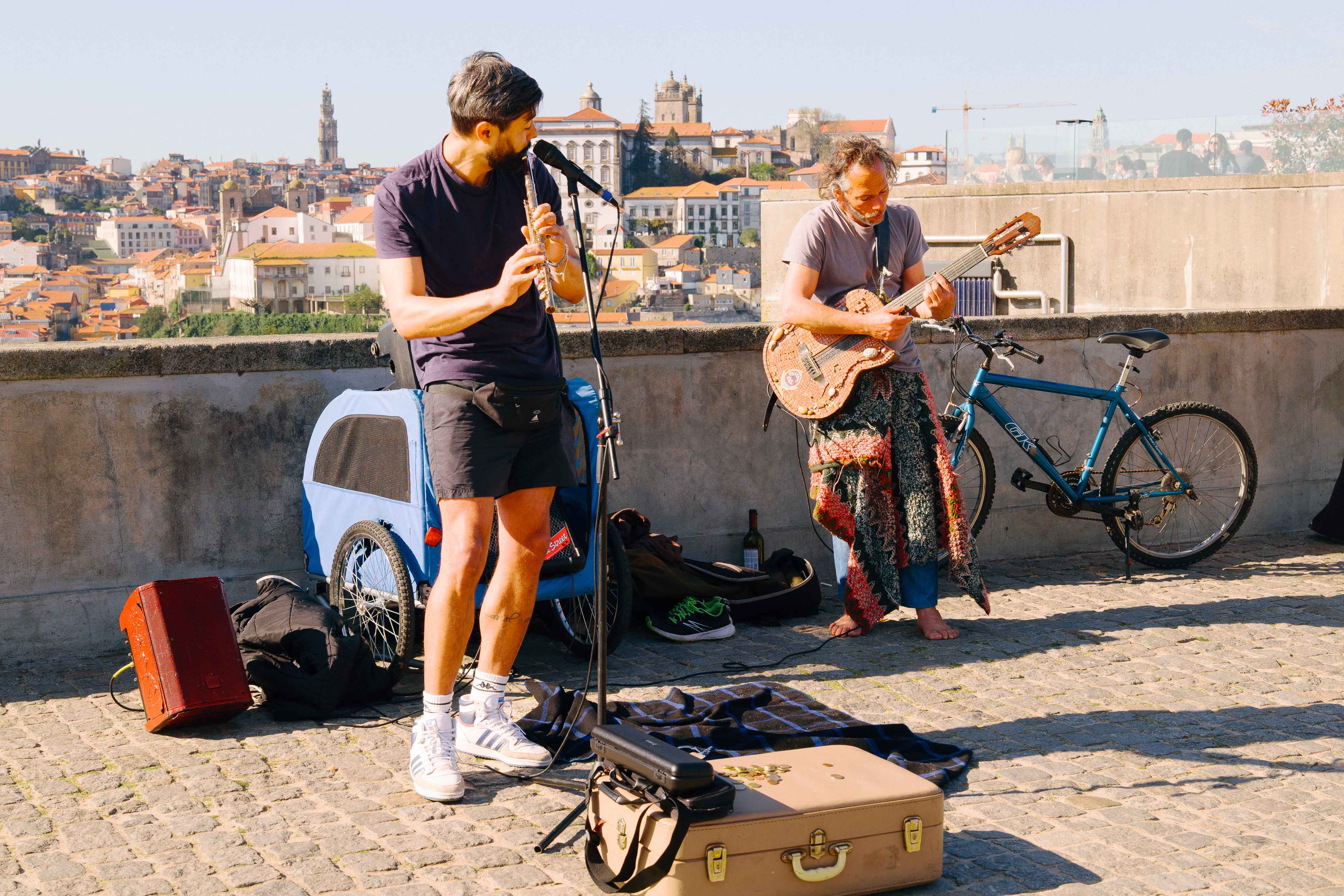 Two street musicians performing on a Porto viewpoint, one playing a flute and the other a guitar, with the city’s historic skyline and tiled rooftops behind them.
