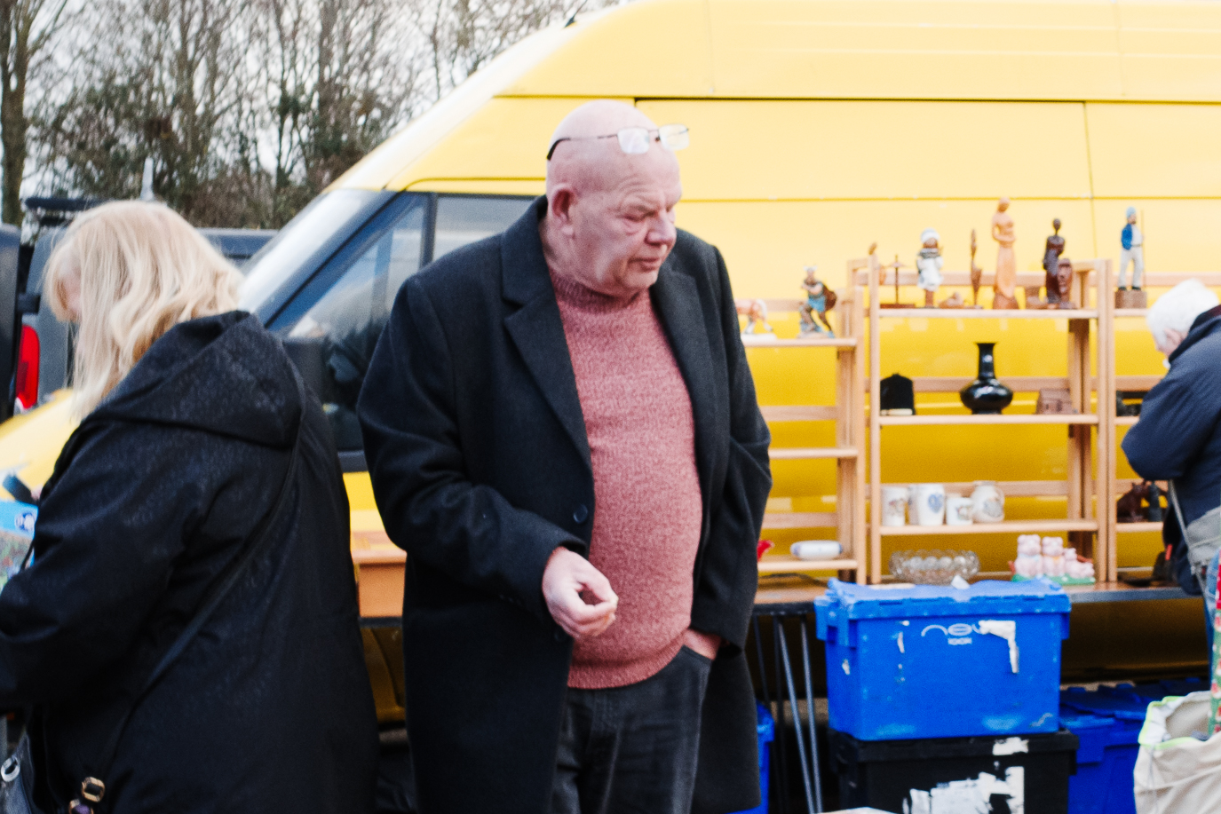 Zuschnitt einer spontanen Szene auf einem Straßenmarkt, die Kisten mit Haushaltsartikeln und Sammlerstücken neben einem leuchtend gelben Van bei natürlichem Tageslicht zeigt. Aufgenommen mit einer Ricoh GR IV. Der Bildzuschnitt zeigt, dass das Gesicht der Person auf dem Bild unscharf ist.