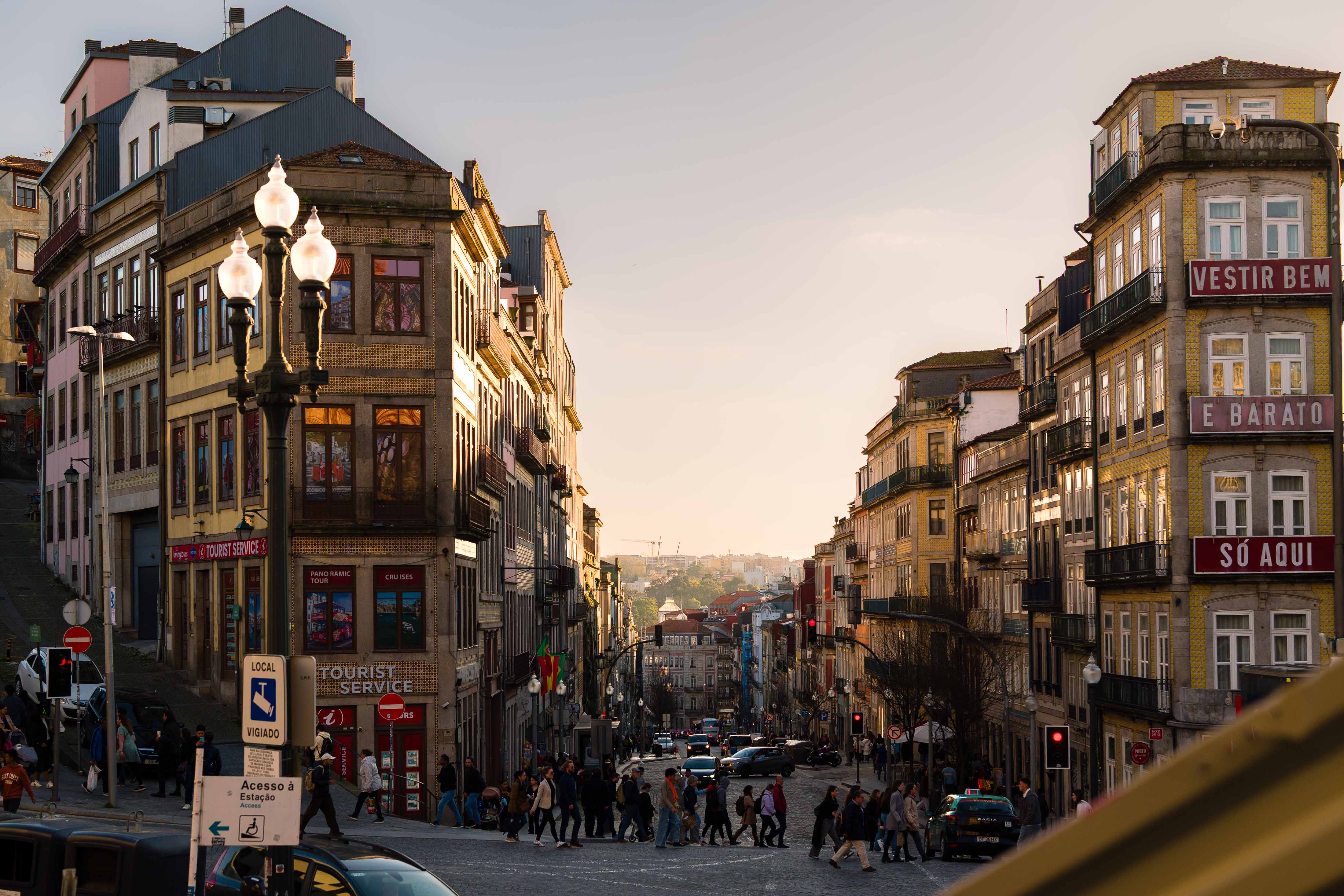Busy Porto street lined with historic buildings and shopfronts, pedestrians crossing the road as the street slopes downhill towards the city centre in warm late-afternoon light.
