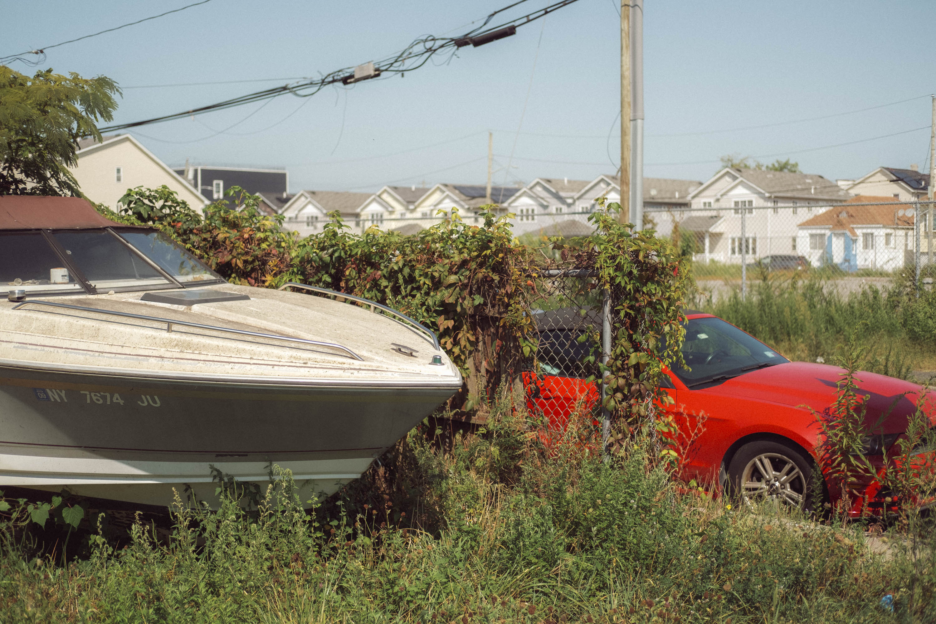 Une voiture rouge et un bateau garé dans un jardin arrière très envahi par la végétation. Photo prise avec le X-Pro 3 par Ian Howorth.