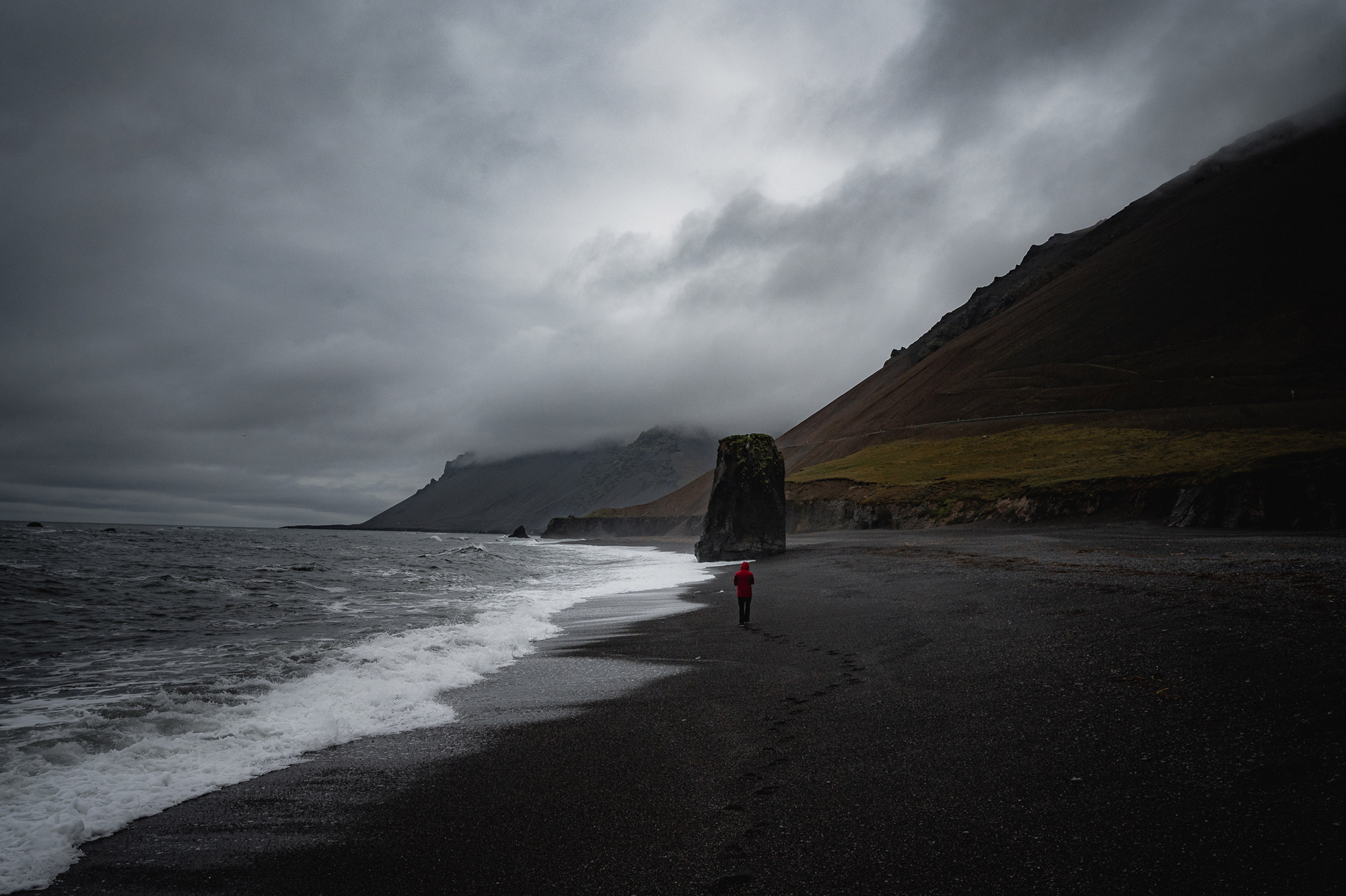 A figure wearing a red coat walks along the coastline in Iceland, with hills and clouds in the background, shot by Paul Watson with a Nikon Z6
