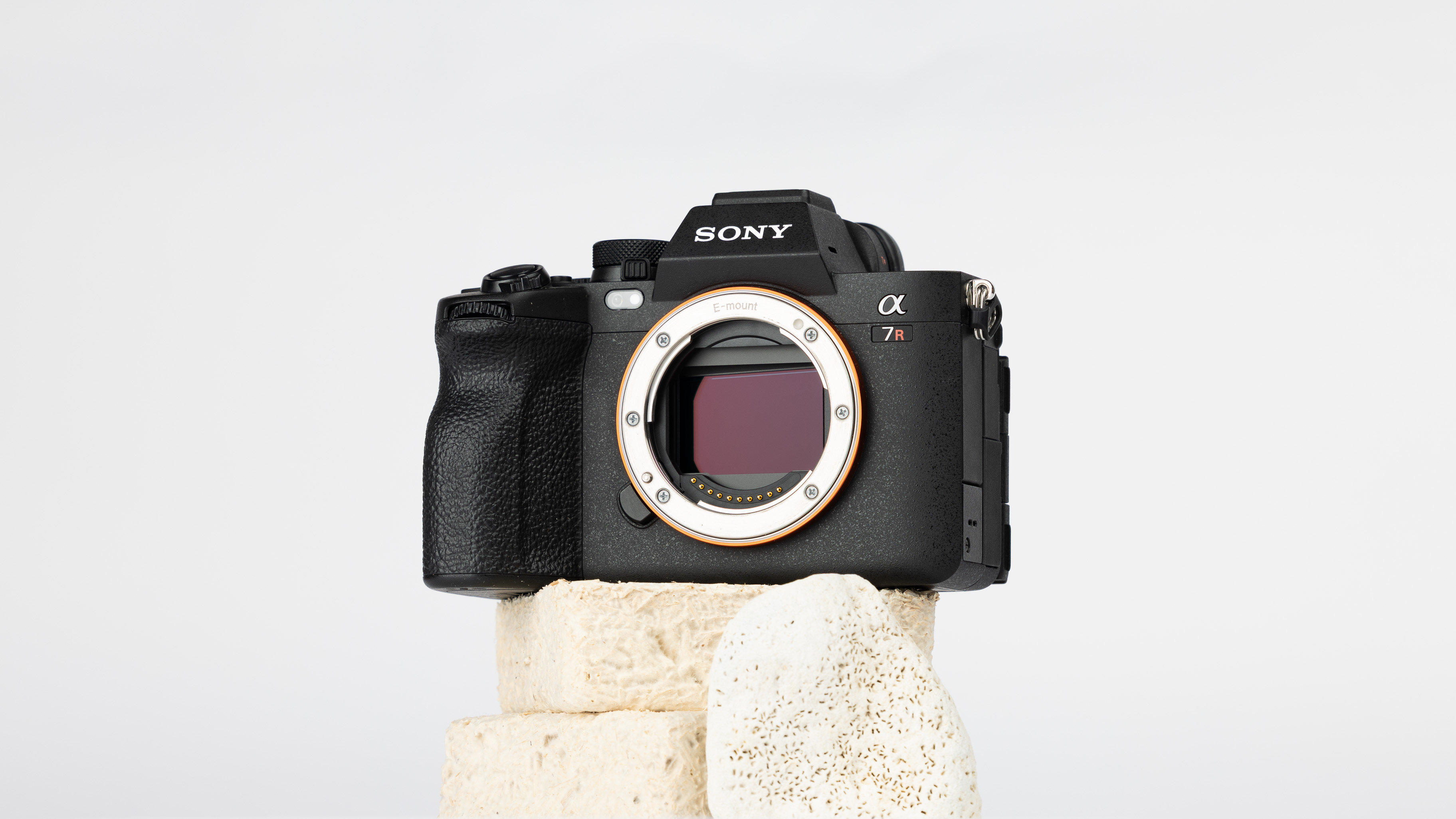 A product shot of a secondhand Sony A7RV camera propped up on a rocky podium on an off white background