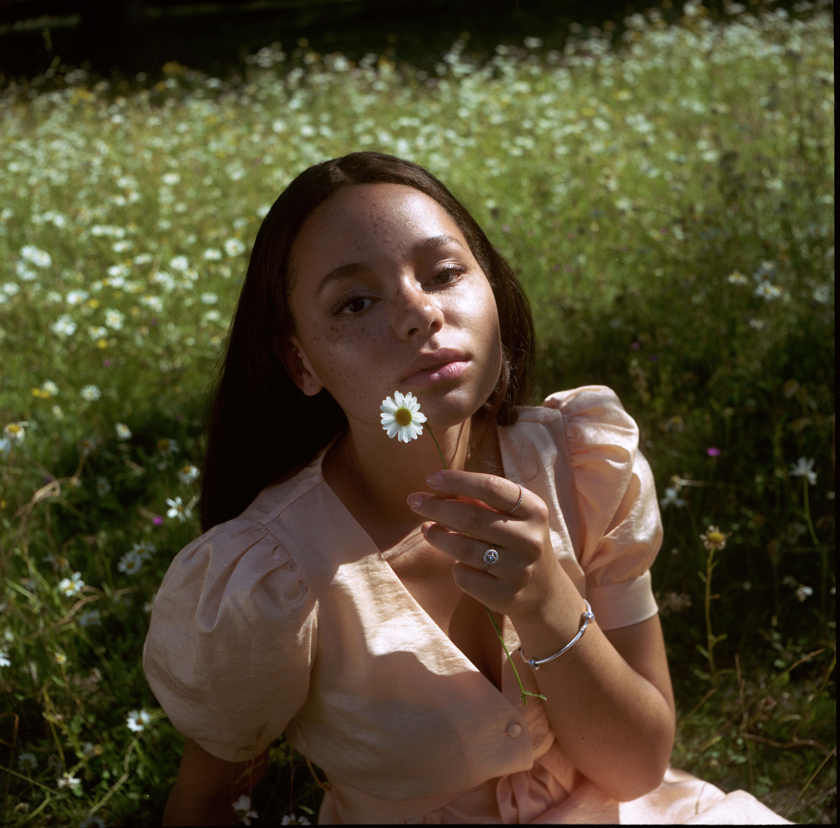 Portrait d'une femme noire portant une robe rose, assise dans l'herbe et tenant une marguerite à la main, réalisé par Samia Rachel.