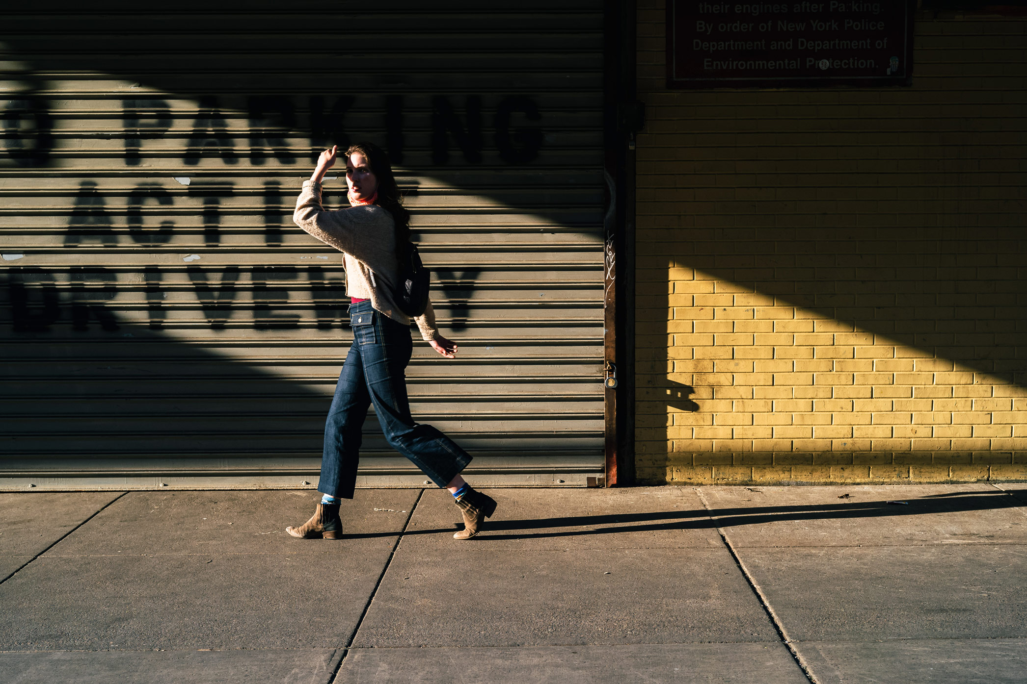 Persoon loopt over stoep voor garage en gele bakstenen muur in New York City en beschermt zijn gezicht tegen de zon, gefotografeerd door Manuel Pena.