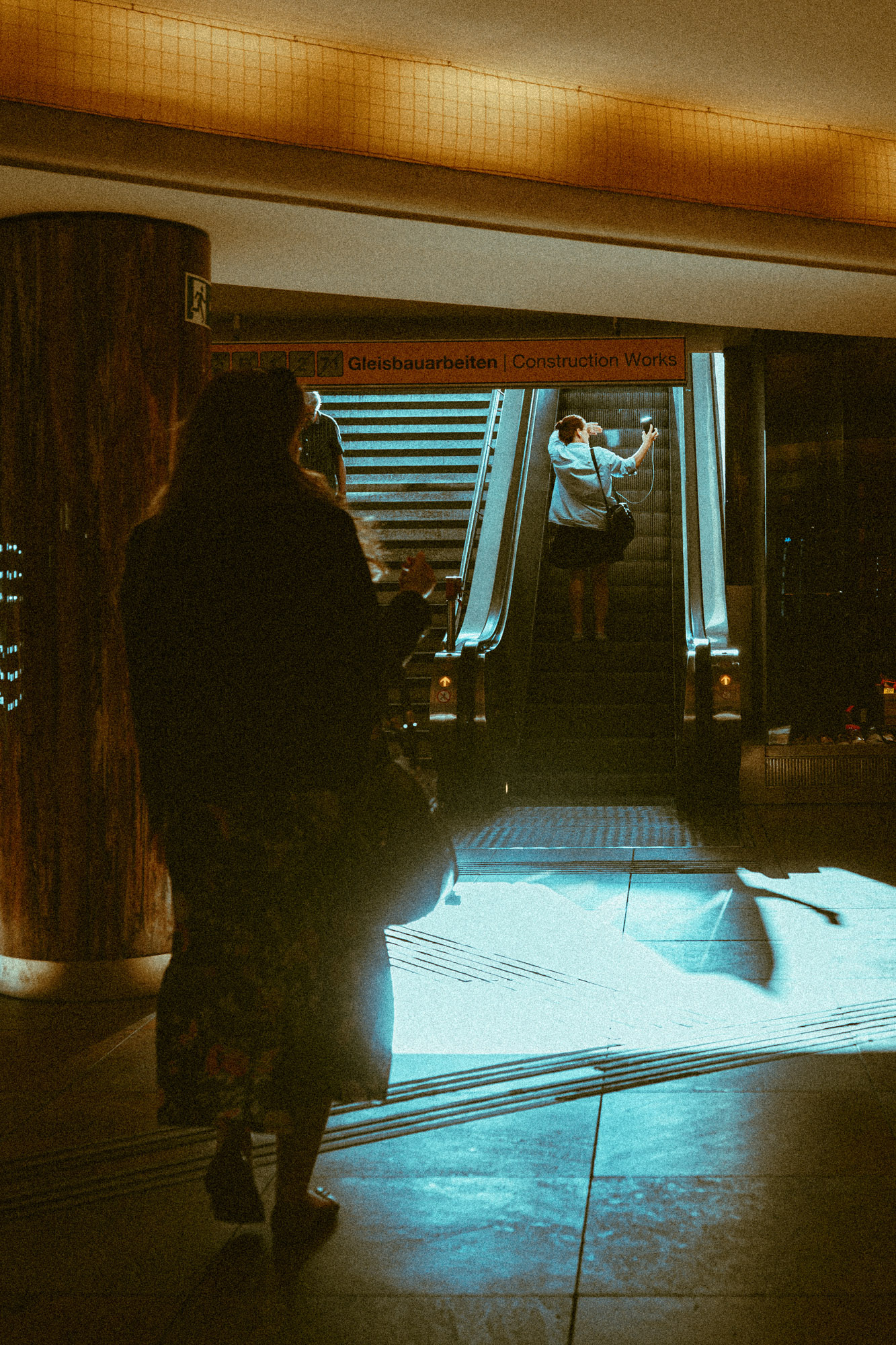 Stimmungsvolles Foto in einer Wiener U-Bahn-Station. Im Vordergrund eine Person, die an einer Säule vorbei in Richtung Rolltreppe zur Straße läuft. Auf der Rolltreppe fährt eine Person nach oben, während eine weitere Person über die daneben liegende Stiege den U-Bahnhof betritt. Von draußen fällt helles Licht auf den Boden der U-Bahn-Station und erzeugt starke Kontraste zwischen warmen und kühlen Farbtönen. Aufgenommen von Dhan Limwattana mit einer Nikon Z5 II.