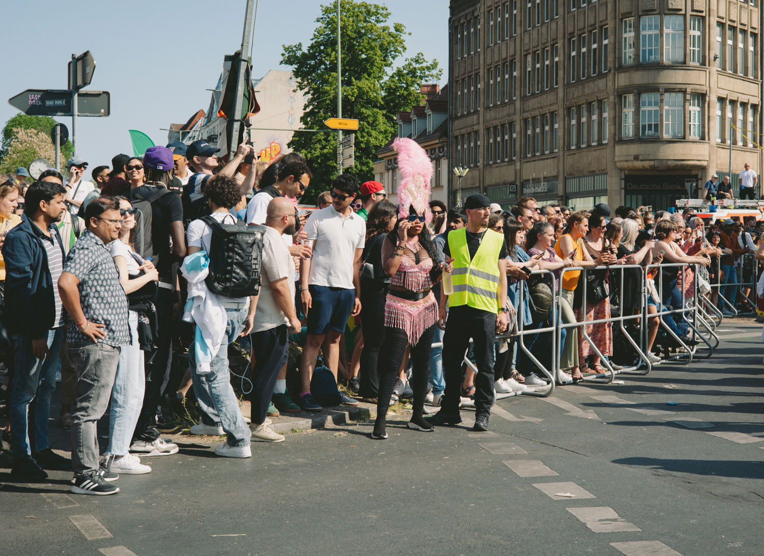 Crowd waiting at a barrier, security person wearing yellow waistcoats and person in a pink costume with feather decoration in the focus of the picture, shot by Stefanie Schmid Rincon with the Canon EOS R6