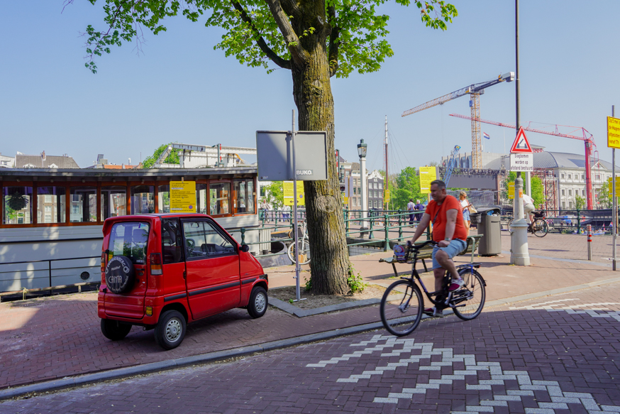 Een levendig Amsterdams straatbeeld met een fietser die langs een rode Canta rijdt bij de gracht, omlijst door lentegroene bomen en stadse details, gefotografeerd door Louise Kluit