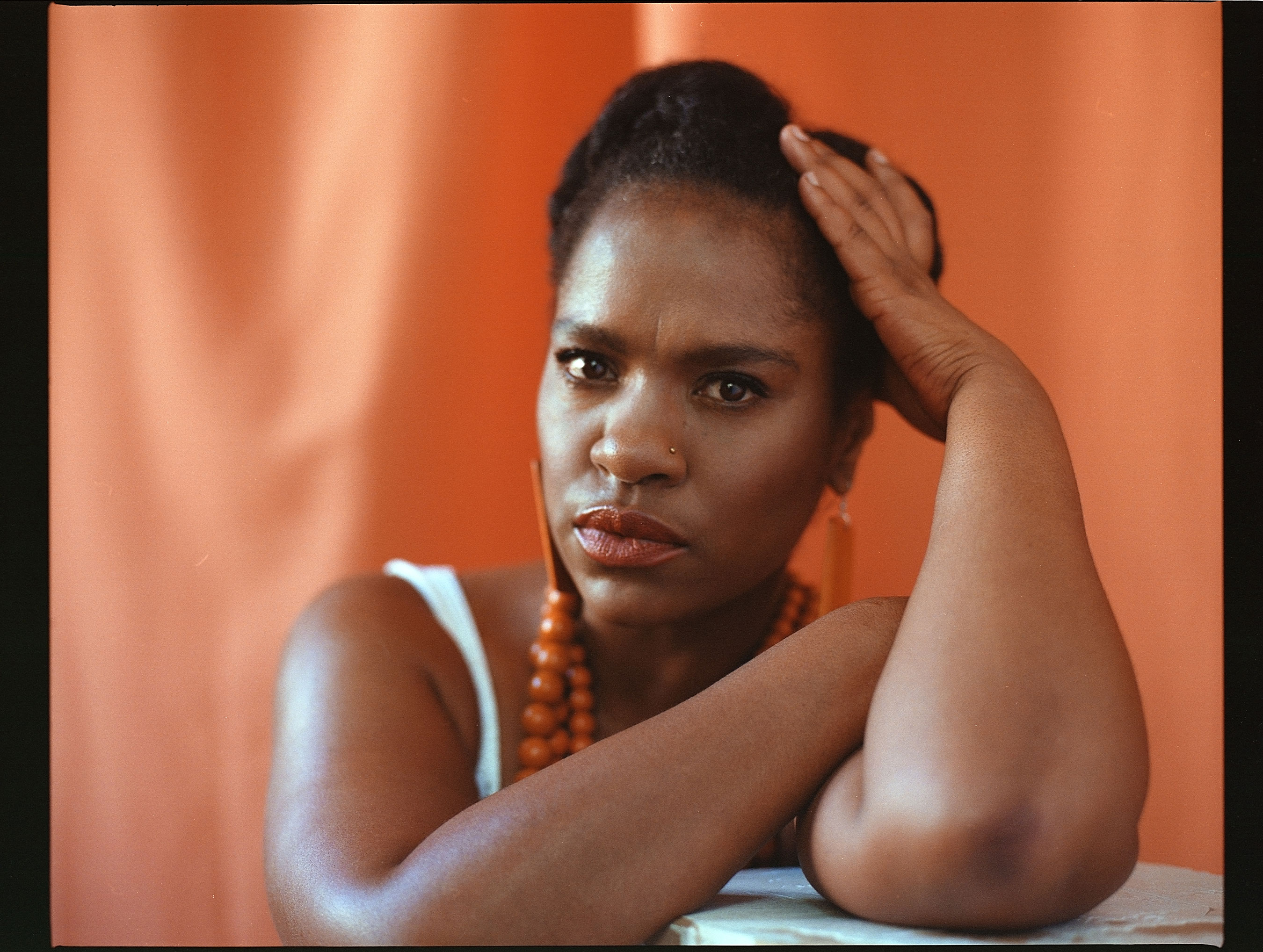 Portrait of a Black woman resting her arms on a pedestal, supporting her head with her left hand and looking seriously into the camera, against an orange background, shot by Samia Rachel
