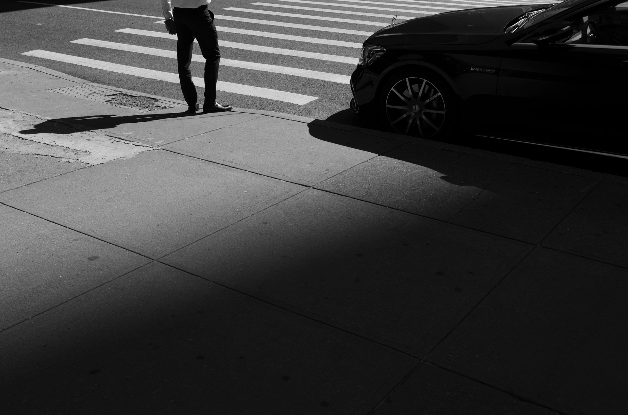 A black-and-white photo of a person standing on the sidewalk by a car