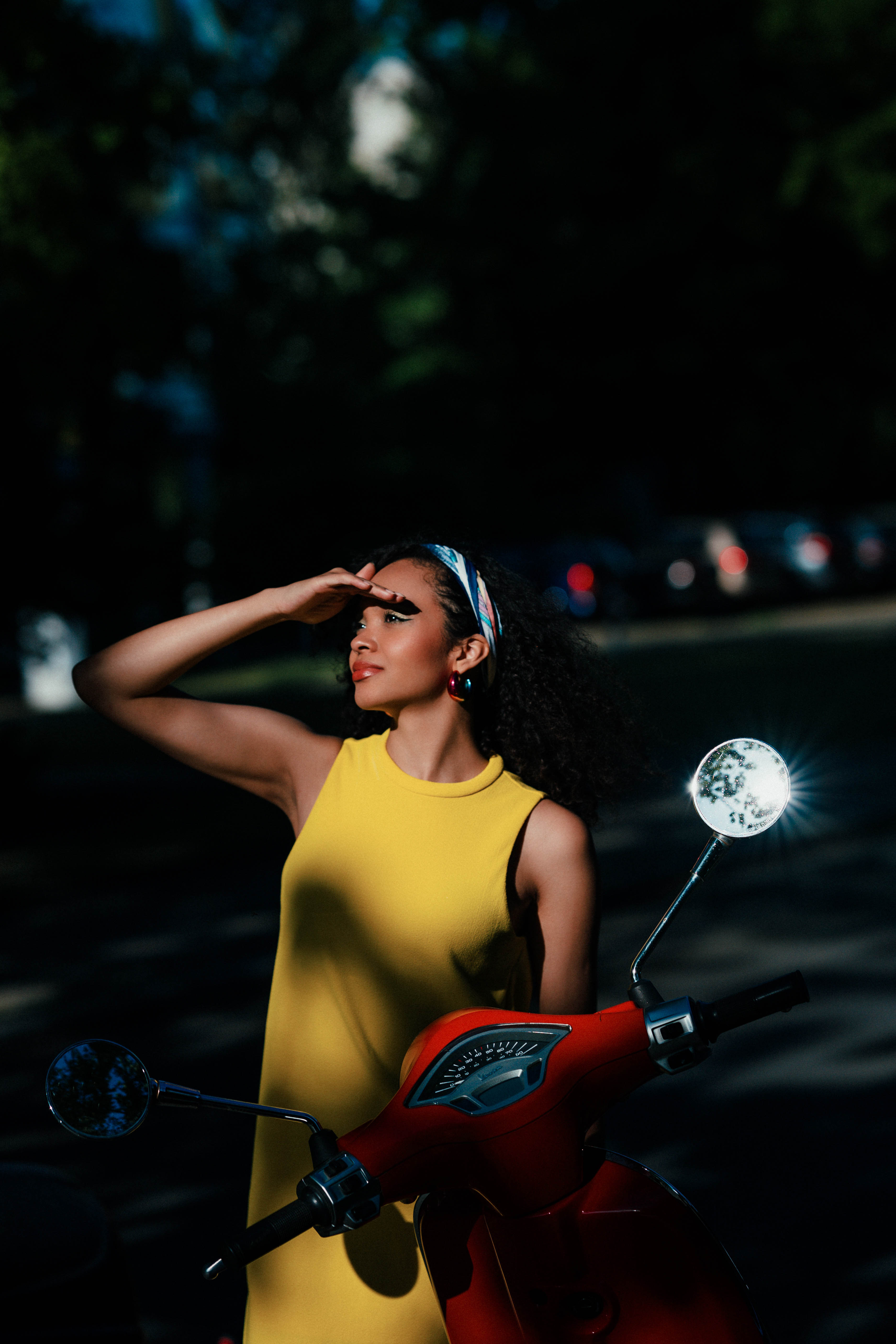 Outdoor portrait of a model in a yellow dress posing with a classic red scooter. Sunlight and shadows create a dramatic effect. Shot by Alina Rudya on a Nikon Z6 III.