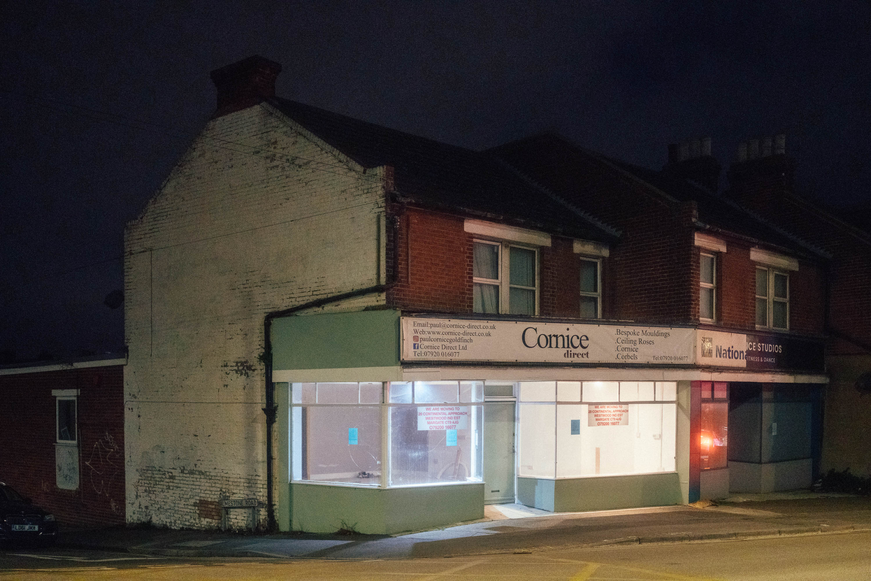 Ein von Straßenlaternen beleuchteter ehemaliger Späti in einer ruhigen Wohnstraße bei Nacht. Die Schaufenster sind beleuchtet und darüber befinden sich ein Vintage-Schild mit der Aufschrift „Cornice Direct“ und eine Backsteinfassade. Aufgenommen von Connor Redmond mit einer Sony RX1R III.