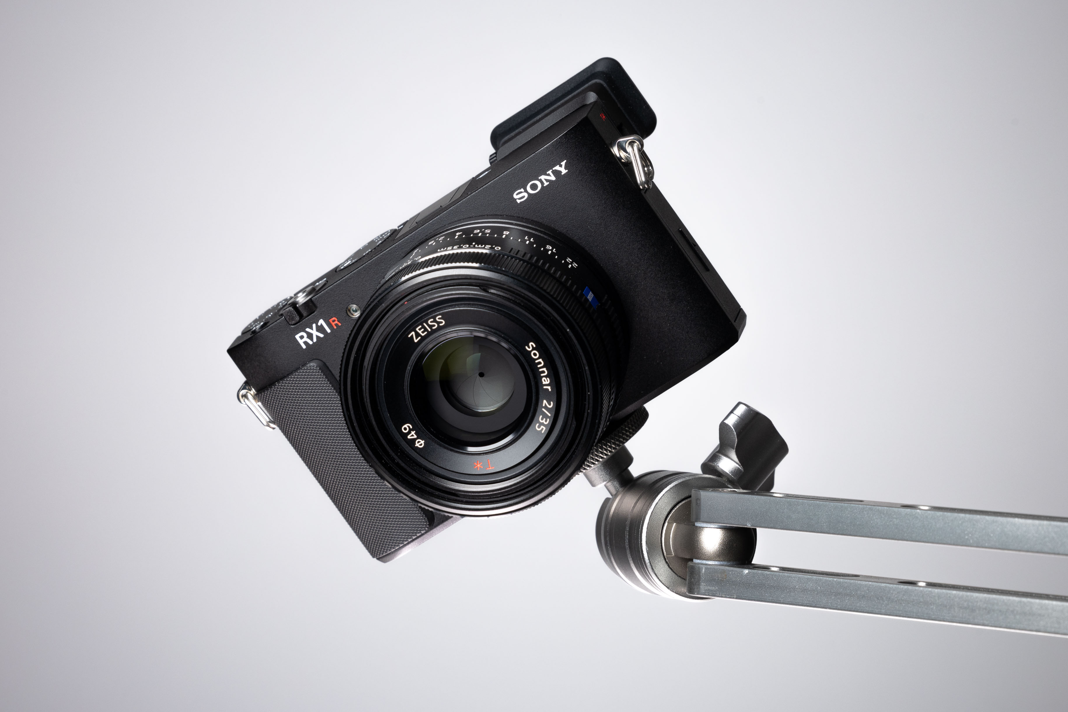 Frontale Studioaufnahme einer schräg auf einem Metallarm montierten Sony RX1R III, die das Zeiss Sonnar T* 35mm F2-Objektiv, den strukturierten Griff und das kompakte Vollformatgehäuse auf weißem Hintergrund zeigt.