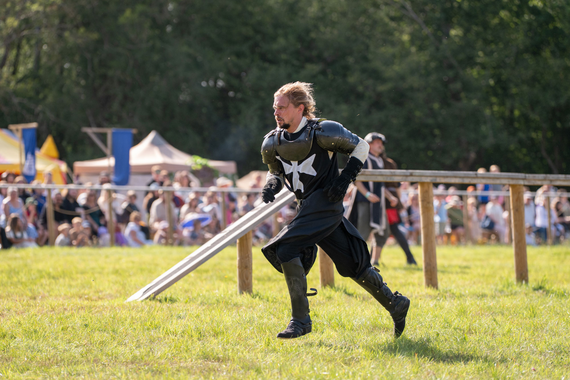 A person in a knight’s costume without a helmet runs from a staged battle. They are mid-sprint. A crowd are in the background, out of focus.