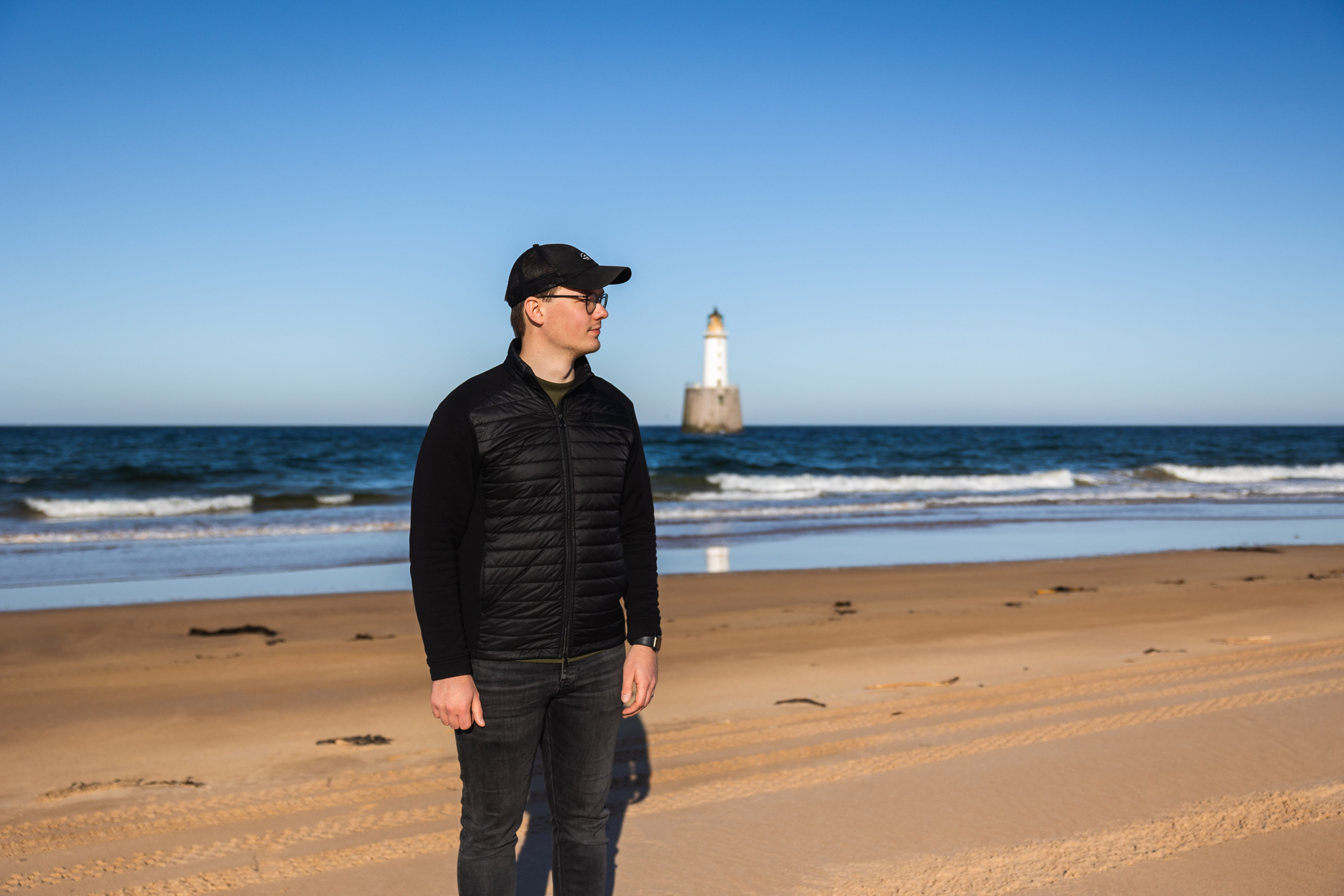 a man wearing a hat standing on a beach with the sea and a lighthouse in the background