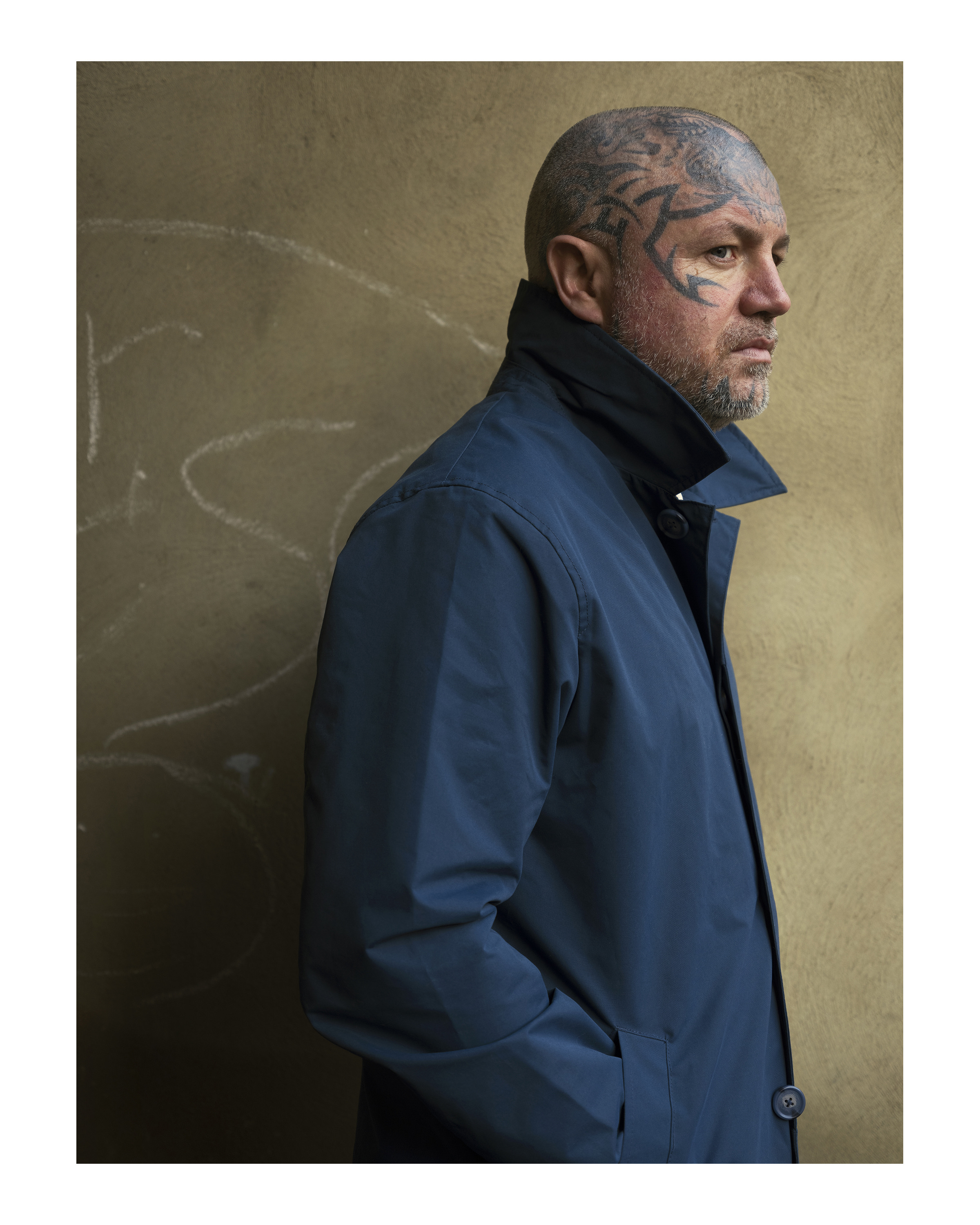 A portrait of a tattooed model looking to the right, wearing a blue coat, standing in front of a wall, photo by Simon Murphy with the Fujifilm GFX 50S