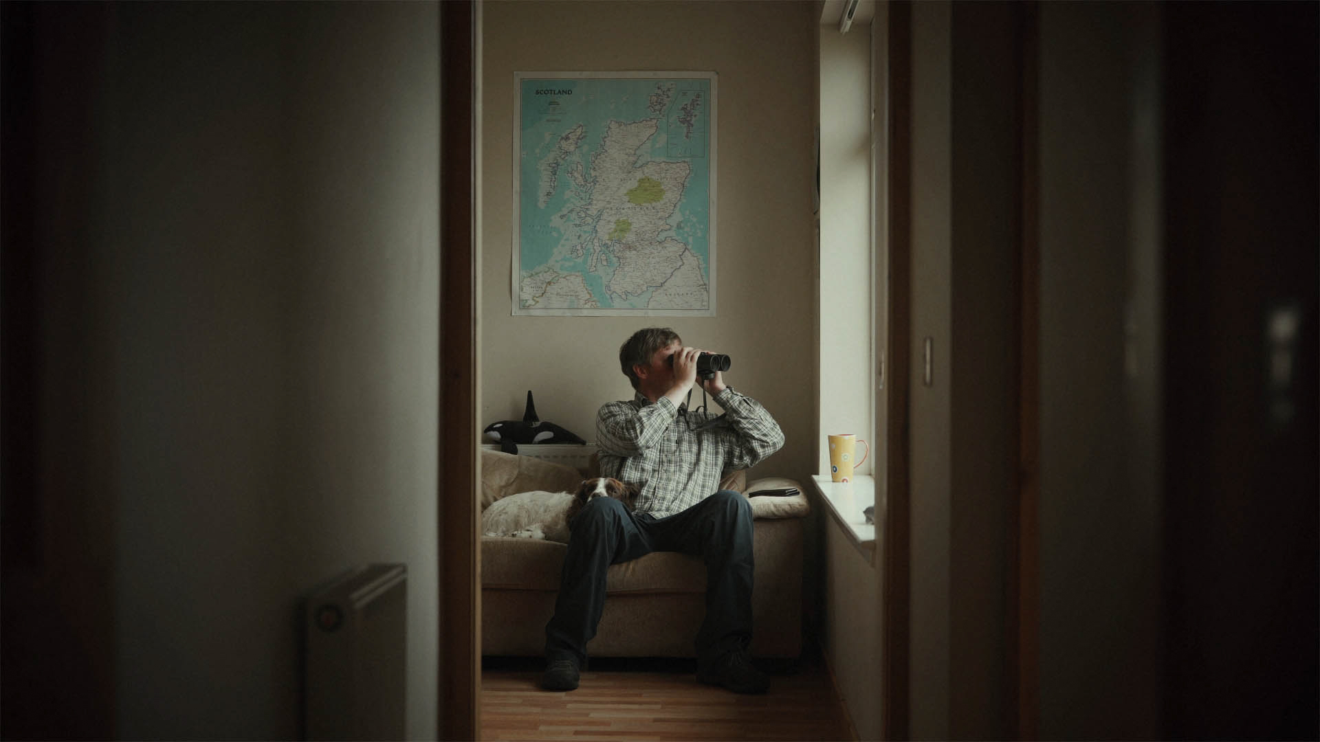 Steve Truluck looking through binoculars by a window below a map of Scotland, a still taken from the short film Truluck by Matt Cannon and Jake Smallwood