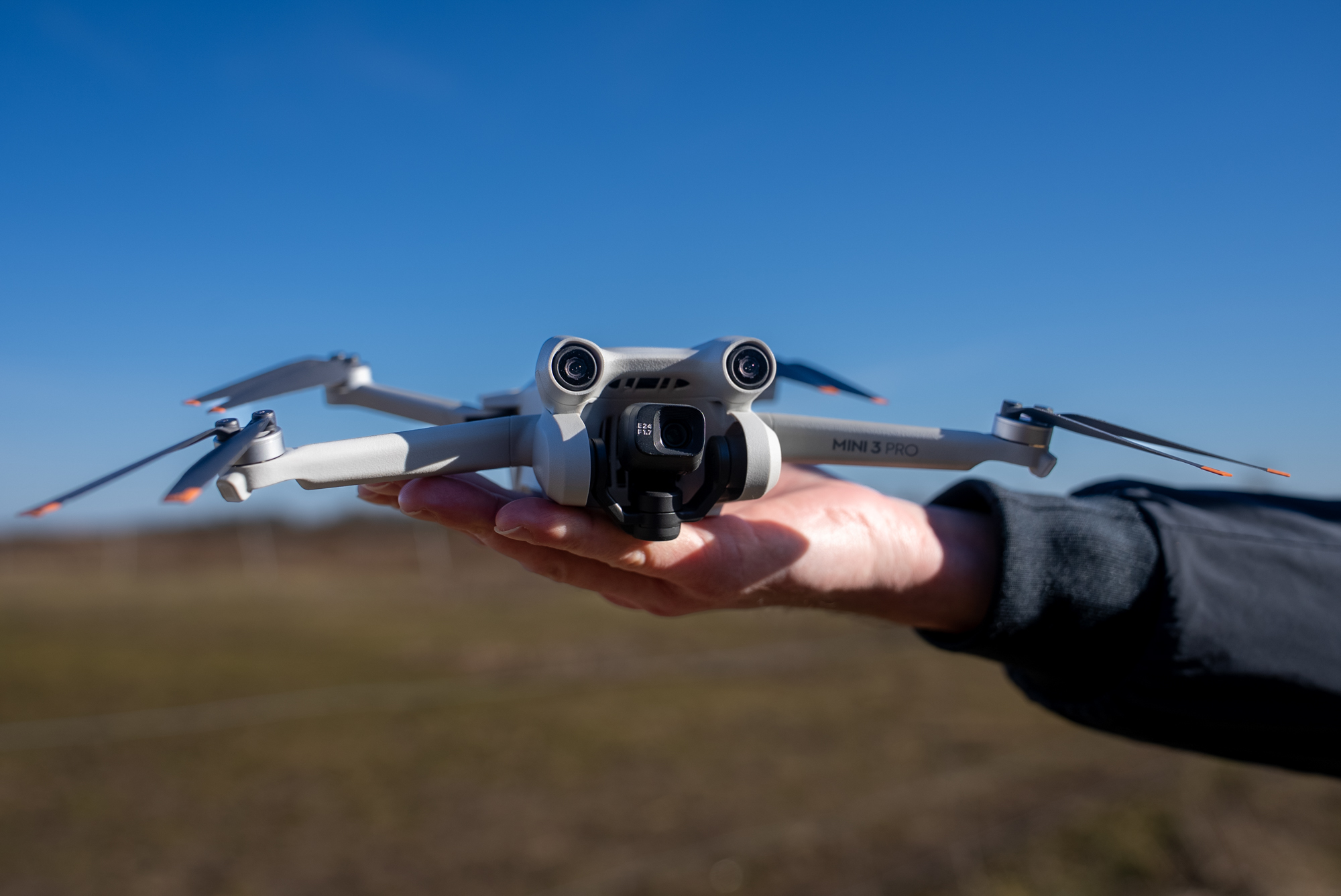 Un compatto drone con fotocamera DJI Mini 3 Pro poggia pronto al decollo su una mano tesa, davanti a un cielo azzurro.