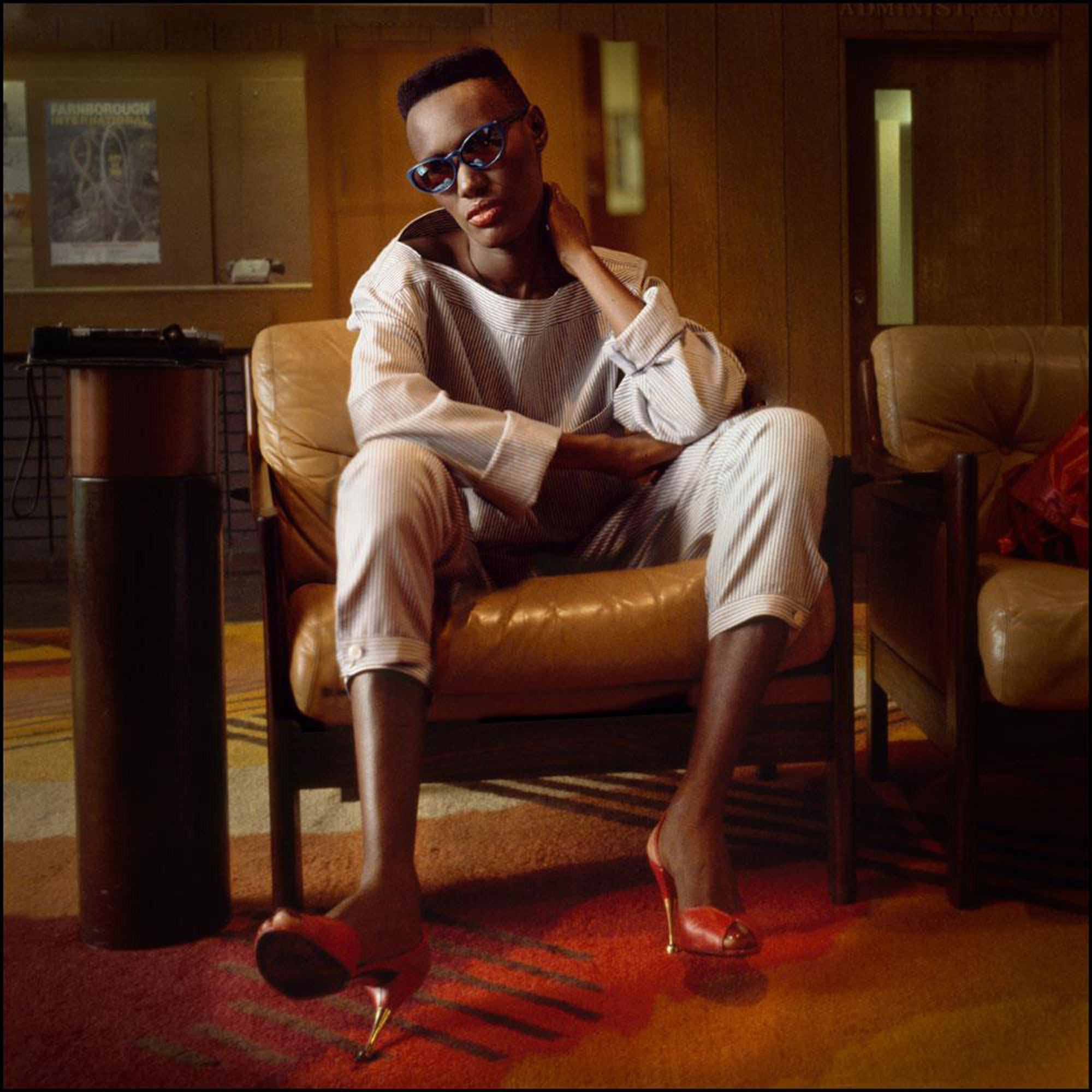 Grace Jones mit Sonnenbrille und roten Schuhen auf einem braunen Lederstuhl sitzend, aufgenommen von Jill Furmanovsky