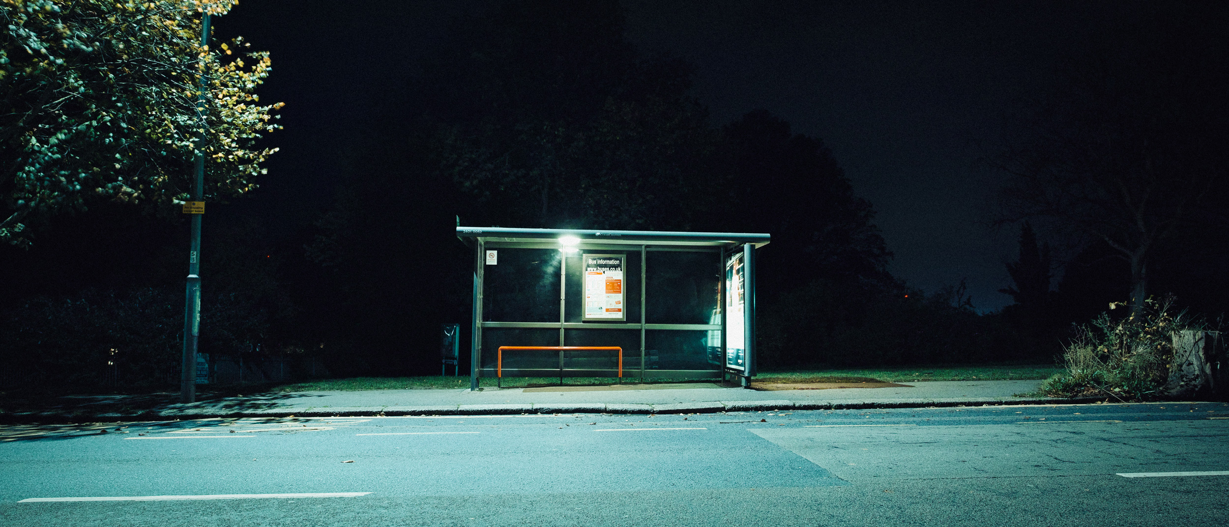 Bushaltestelle mit Glasscheiben und einer orangefarbenen Bank am Straßenrand bei Nacht, aufgenommen mit einer Sigma DP0 Quattro von Jakub Golis