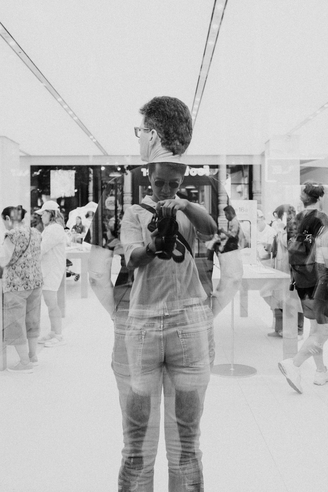 Schwarz-Weiß-Aufnahme in Wien mit Spiegelungs- oder Doppelbelichtungseffekt: Im Vordergrund ist eine Person mit Kamera zu sehen, deren Spiegelung eine weitere Person in einem hellen Ladengeschäft oder Ausstellungsraum überlagert. Im Hintergrund, mehrere Menschen. Aufgenommen von Dhan Limwattana mit einer Nikon Z5 II.