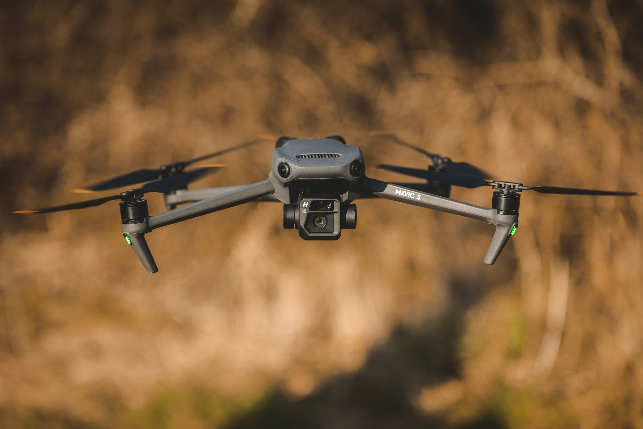 DJI Mavic 3 drone hovering mid-air, showing the front camera system and extended arms against a blurred background.