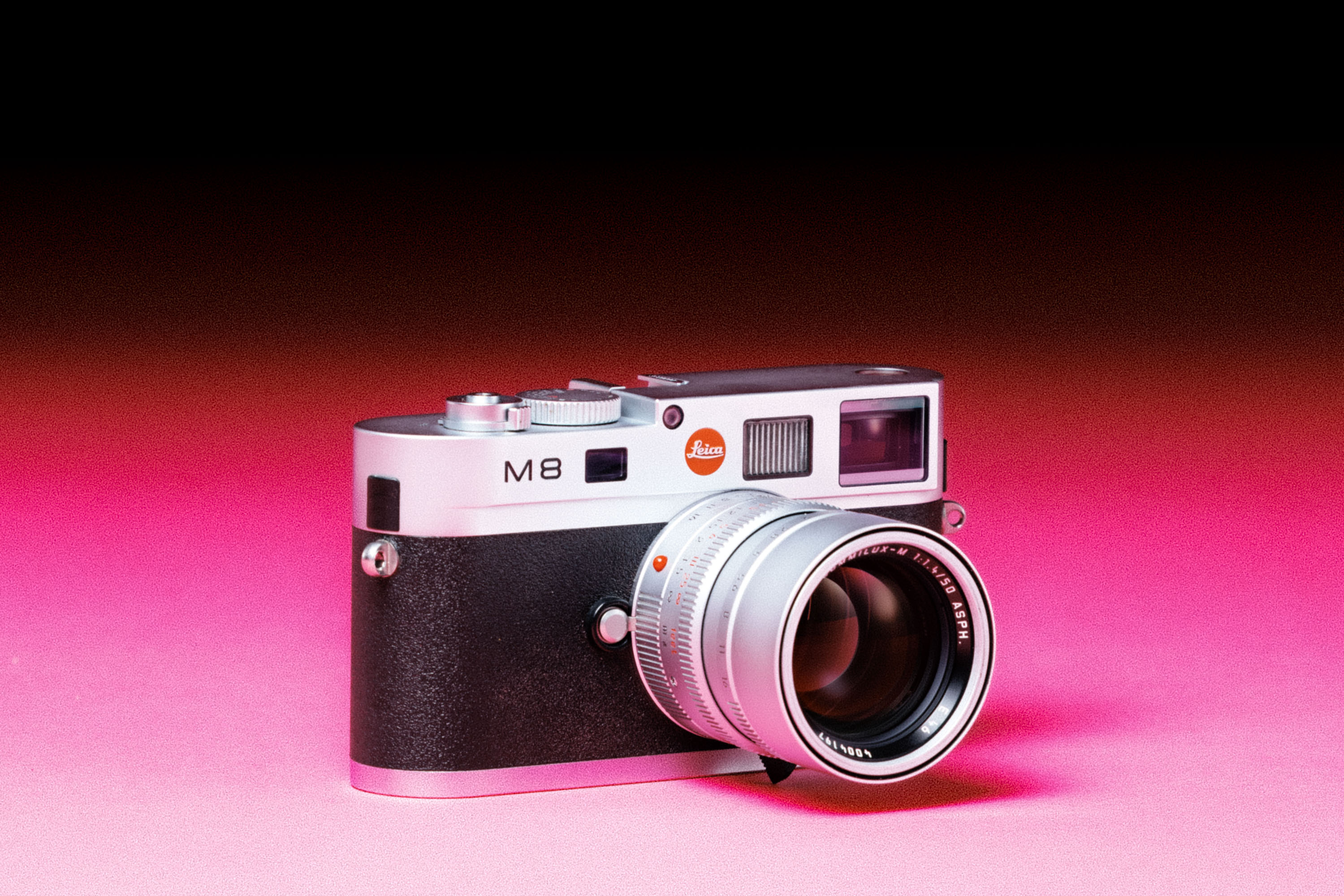 A used Leica M8 camera with a Leica 50mm f/1.4 Summilux-M ASPH lens, with an added film-like grain effect on a graduated pink background. 