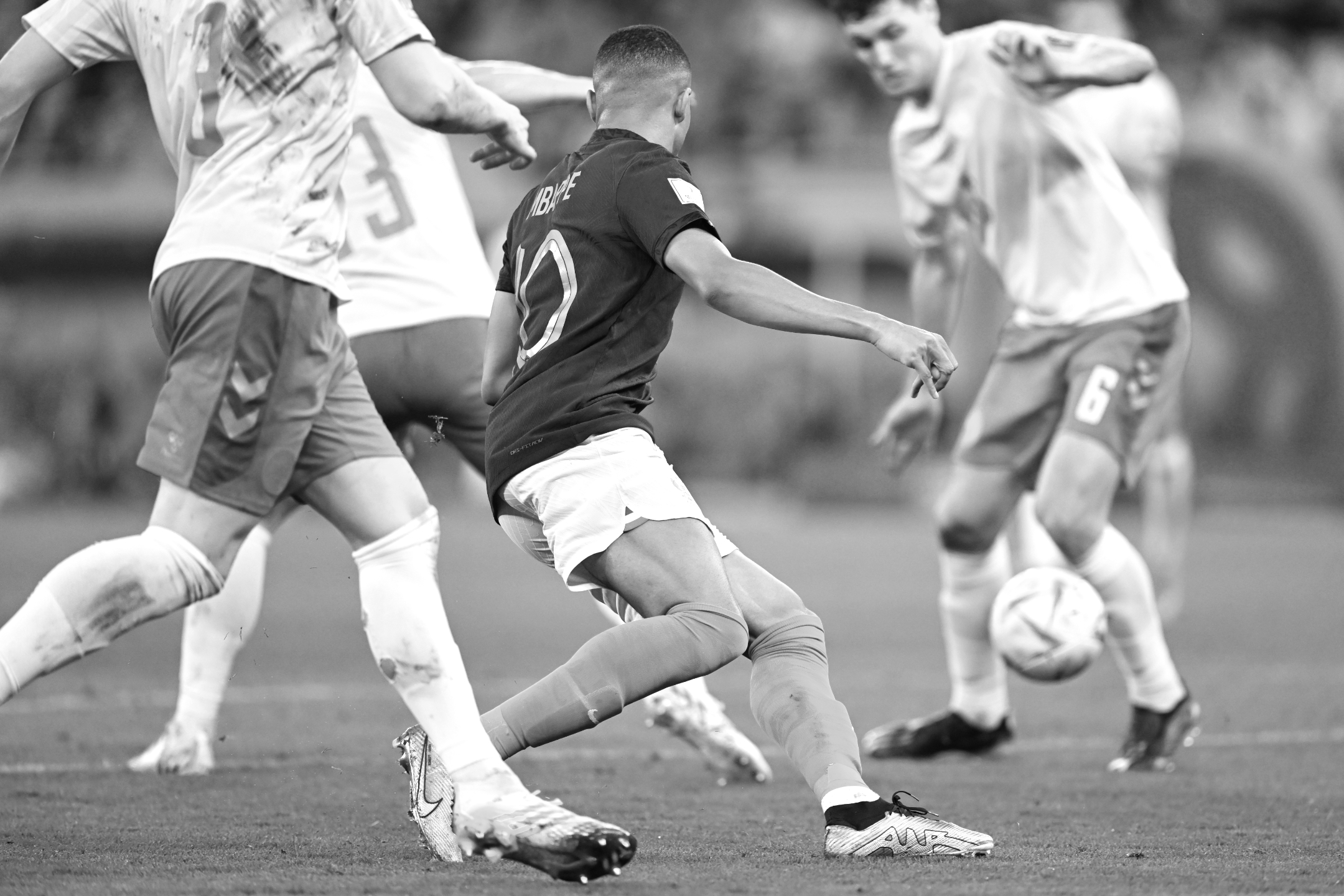 Ein Schwarz-Weiß-Foto des französischen Nationalspielers Kylian Mbappe, der umringt von dänischen Spielern während des 2:1-Sieges seiner Mannschaft bei der Fußball-Weltmeisterschaft 2022, bei dem er zwei Tore erzielte, dem Ball hinterherläuft, aufgenommen von Ryu Voelkel mit einer Nikon Z9.