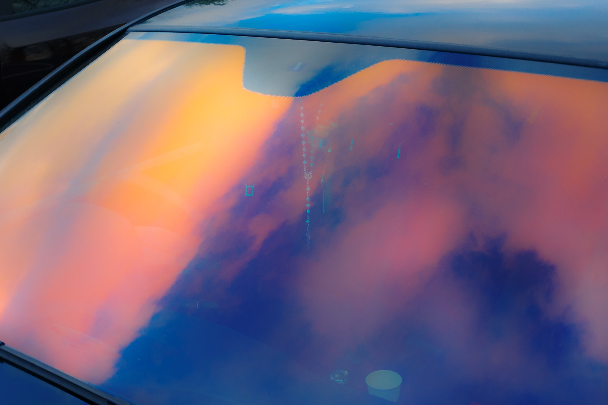 Abstrakte Aufnahme bei natürlichem Licht, auf der sich Wolken in zartem Rosa, Orange und Blau auf der Windschutzscheibe eines Autos spiegeln. Aufgenommen mit einer Ricoh GR IV.