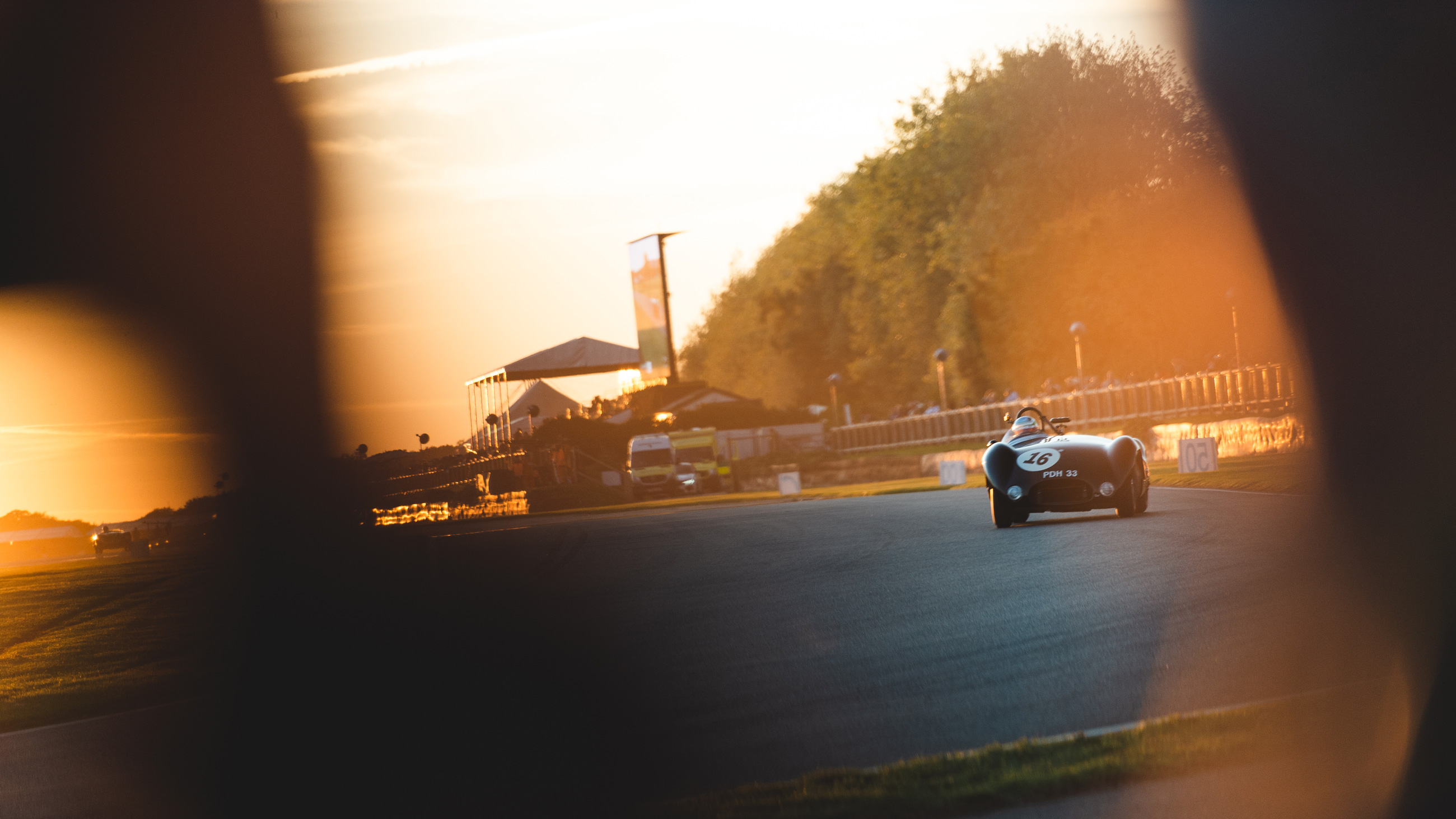 Een wijde opname van een donkere klassieke motorsportauto, genummerd 16, rijdt over een circuit op Goodwood Revival tijdens zonsondergang, gefotografeerd door het raam van een andere auto
